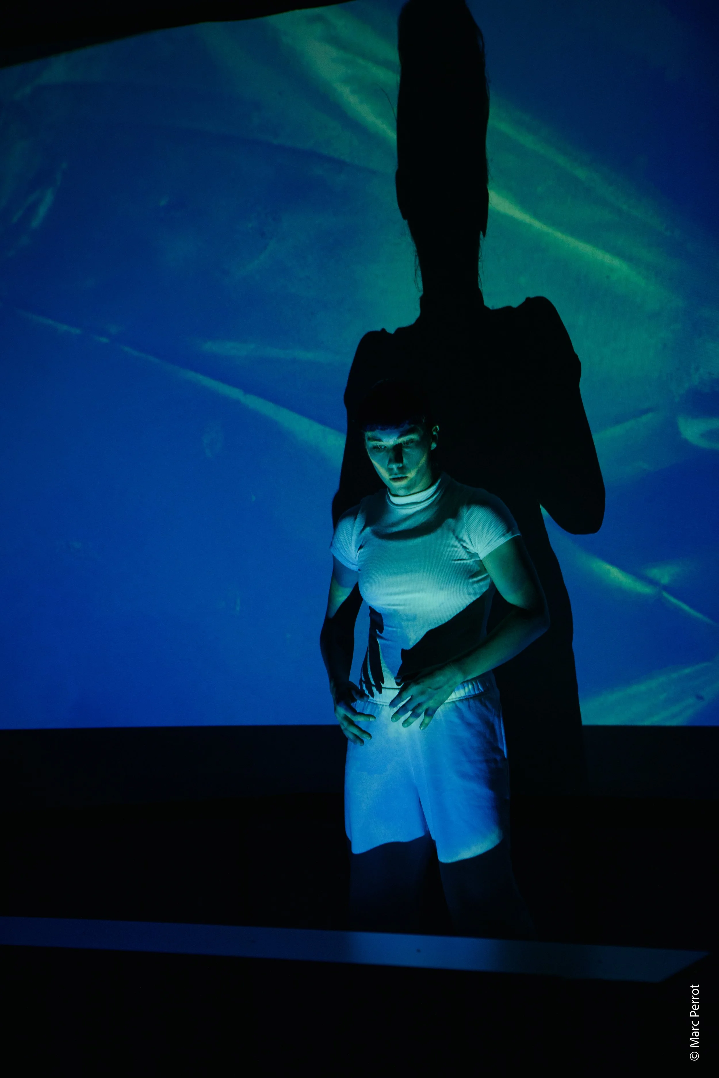 Deux personnes, dont une en blanc, sont dans un environnement sombre avec des projections de lumières bleues et vertes. La personne en blanc regarde vers le bas, la tête légèrement inclinée, tandis que la silhouette derrière est plus grande, avec des