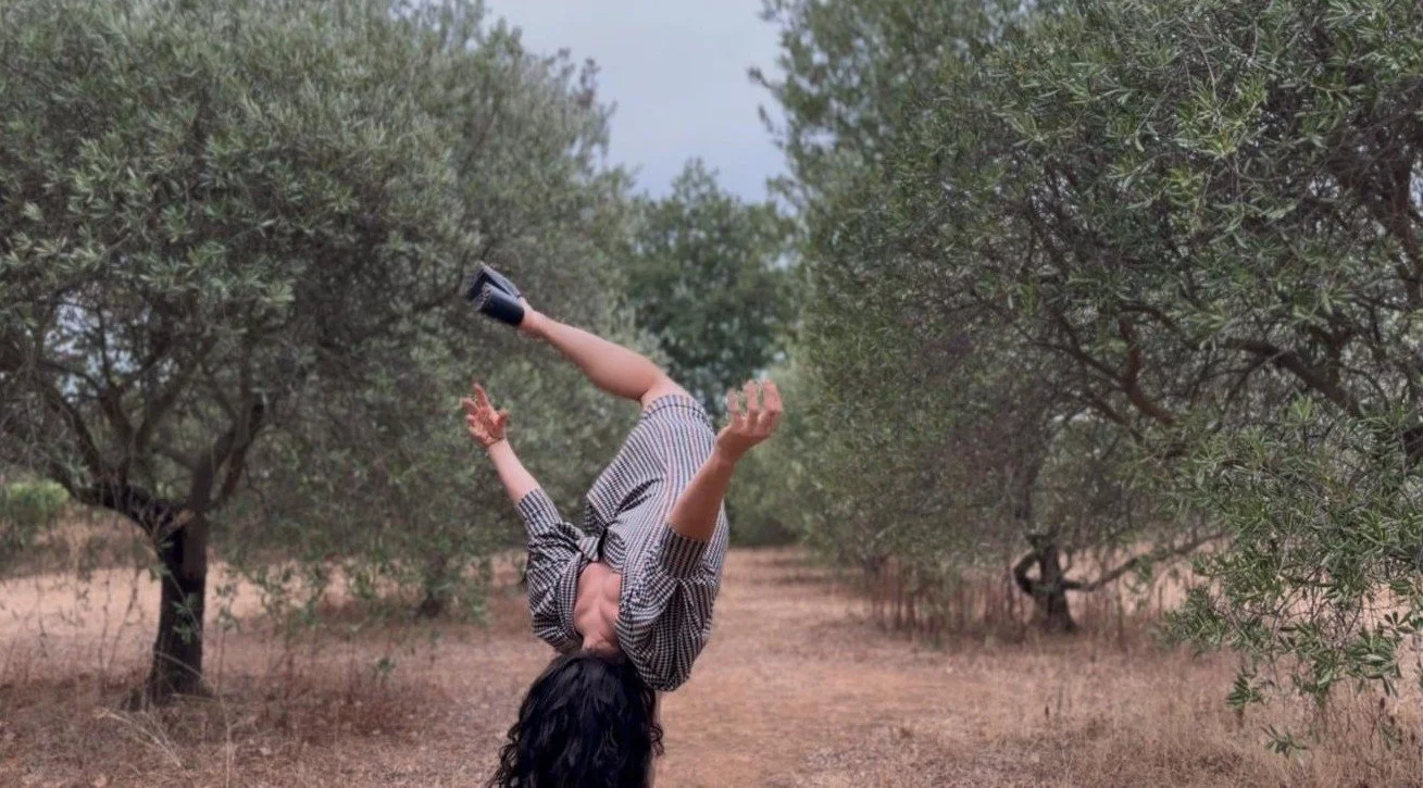 Une personne effectuant une acrobatie en équilibre sur sa tête entre deux rangées d'arbres dans un champ sec.