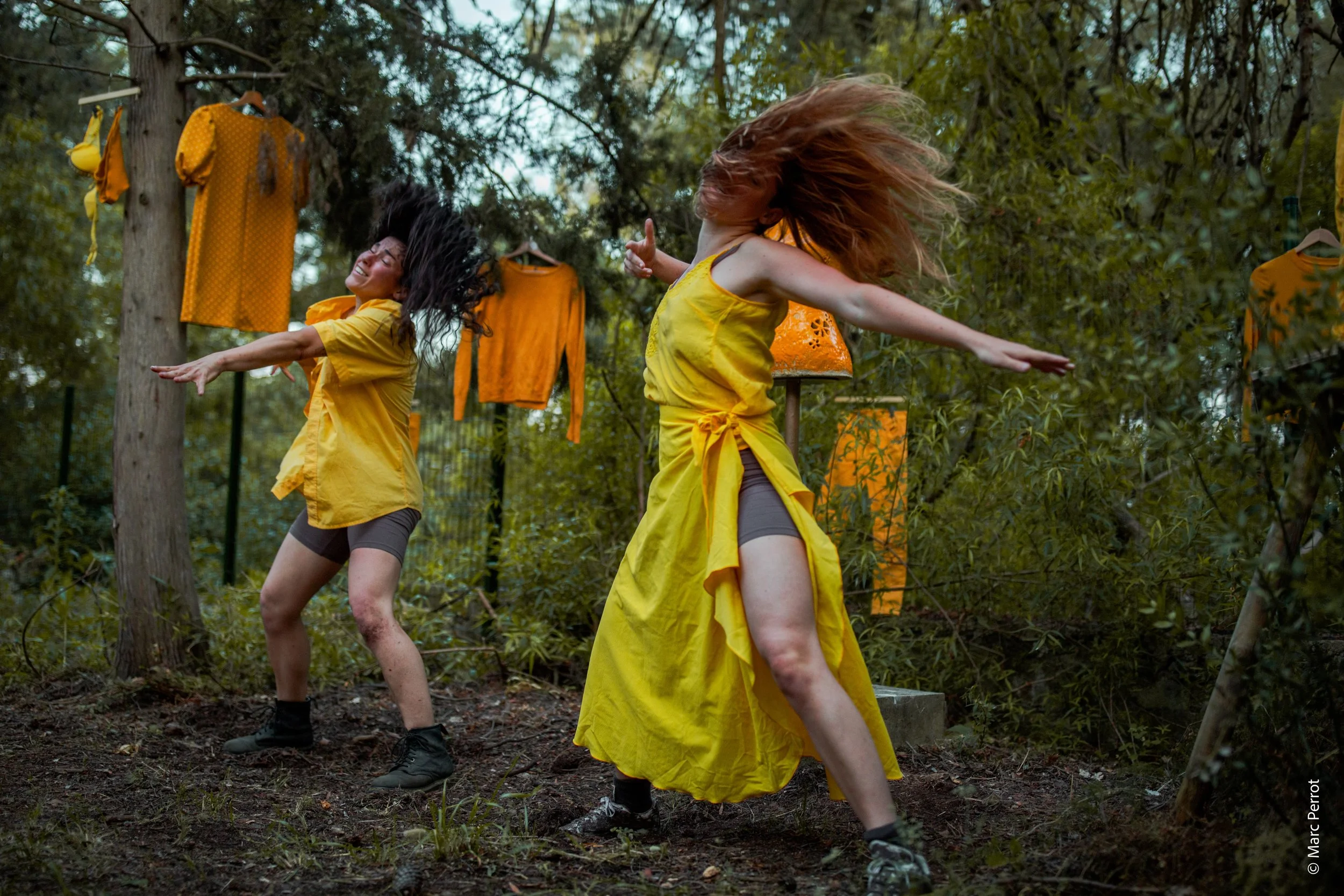 Deux femmes dans un bois, portant toutes deux des vêtements jaunes, dansent ou sautent avec des cheveux en mouvement, entourées de vêtements orange suspendus aux arbres.