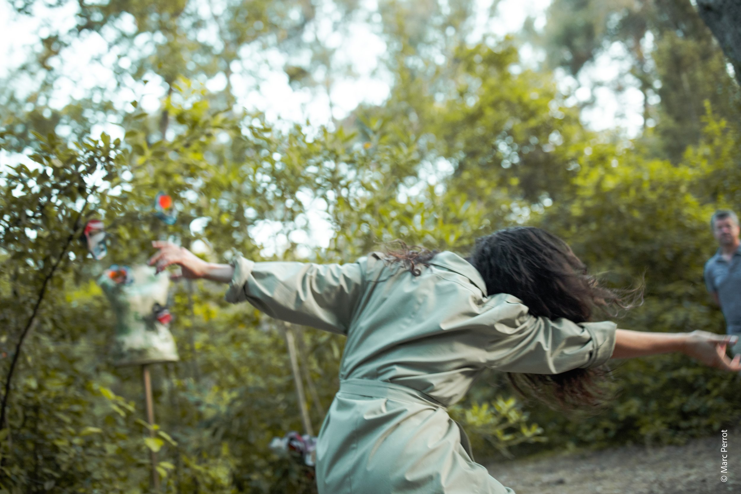 Une personne portant une veste beige lance un bouquet de fleurs en l'air dans une forêt avec d'autres personnes en arrière-plan.
