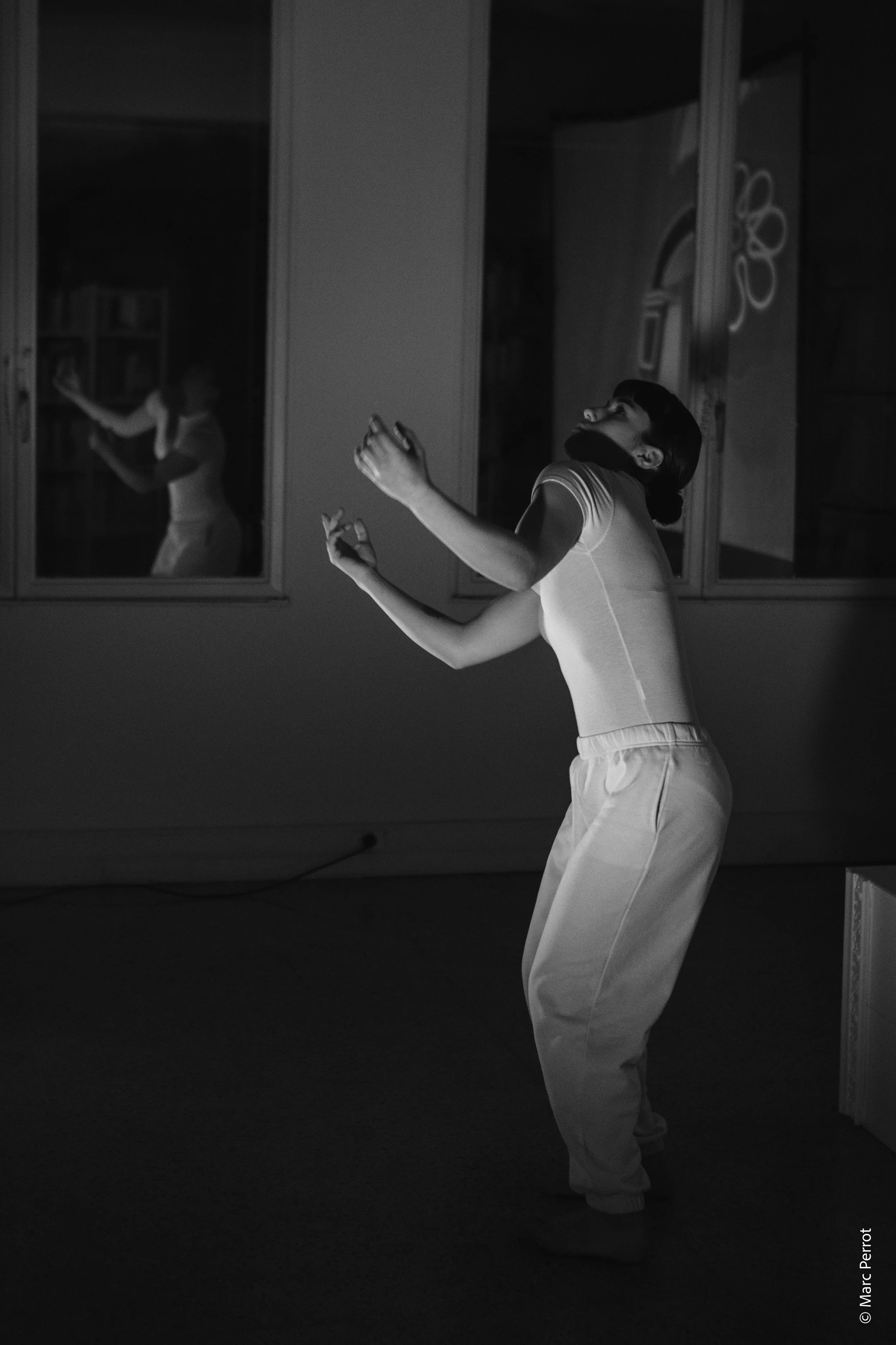 Une personne en tenue sportive effectuant une activité physique dans une pièce sombre. La personne a les bras levés et est vue de profil, avec un reflet d'elle dans une fenêtre ou un miroir derrière elle.