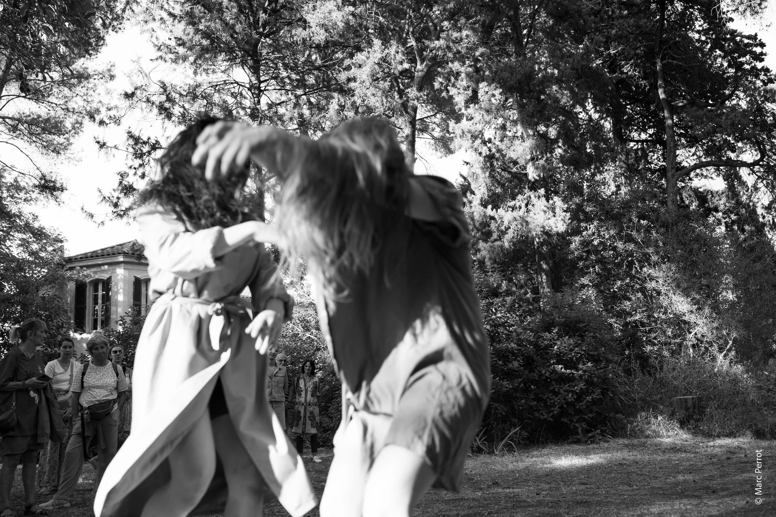 Deux personnes dansent en plein air devant un groupe de spectateurs, avec des arbres en arrière-plan.