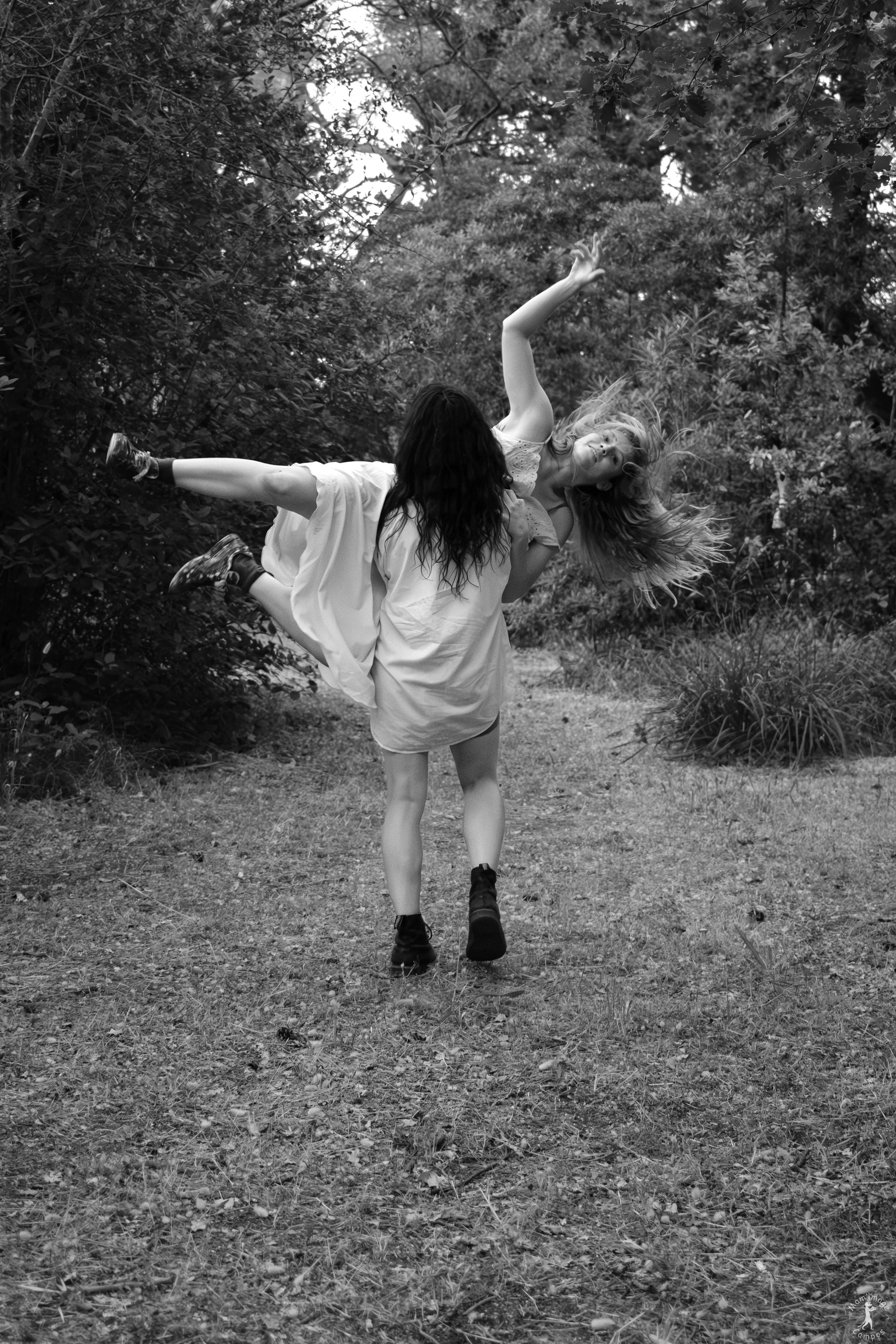 Deux jeunes femmes dans une forêt. L'une d'elles est portée en l'air par l'autre, qui la soutient par la taille. Les femmes portent des vêtements décontractés et semblent en mouvement ou dans une danse.