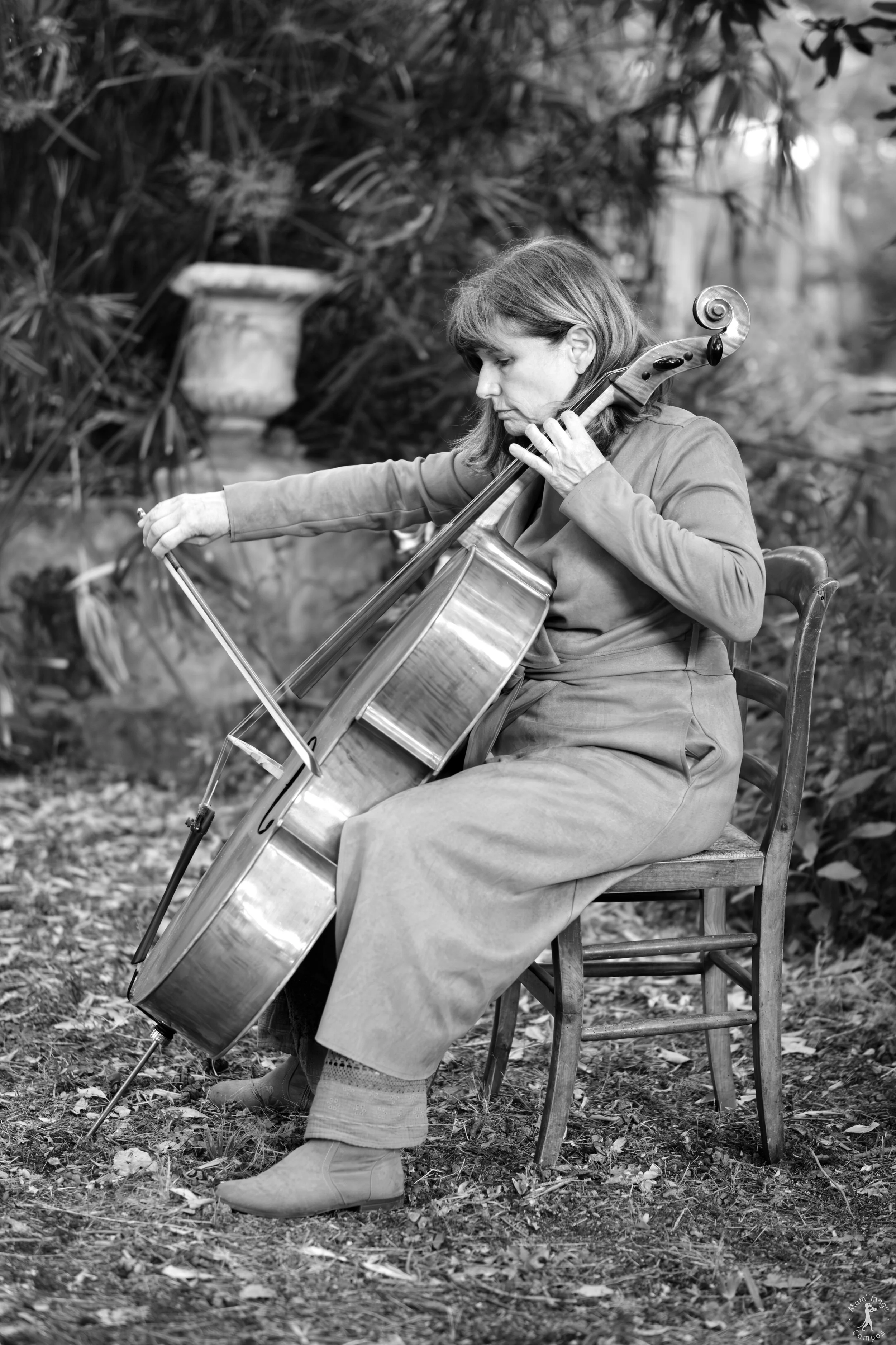 Une femme assise sur une chaise joue du violoncelle dans un jardin en plein air, vêtue d'une robe longue et de bottes, entourée de végétation.
