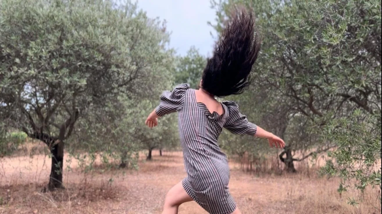 Femme en robe à carreaux courant ou dansant dans une forêt avec des arbres, ses cheveux longs volent dans l'air.