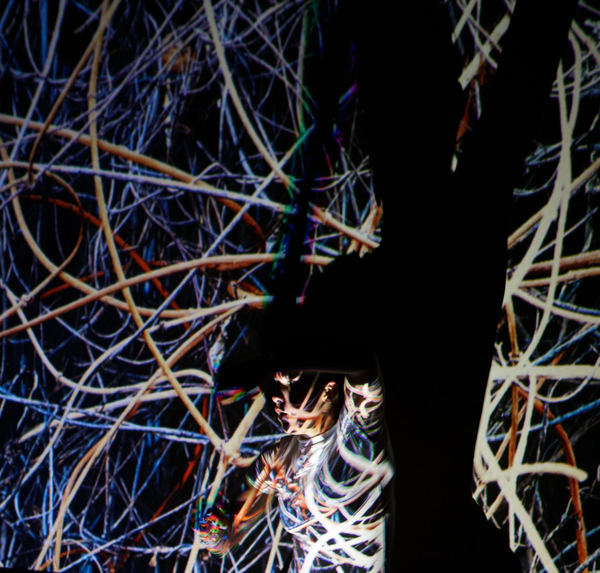Un homme vêtu de vêtements colorés avec un casque à réalité virtuelle. Il est entouré d'une structure composée de branches ou de fils lumineux dans un espace sombre.