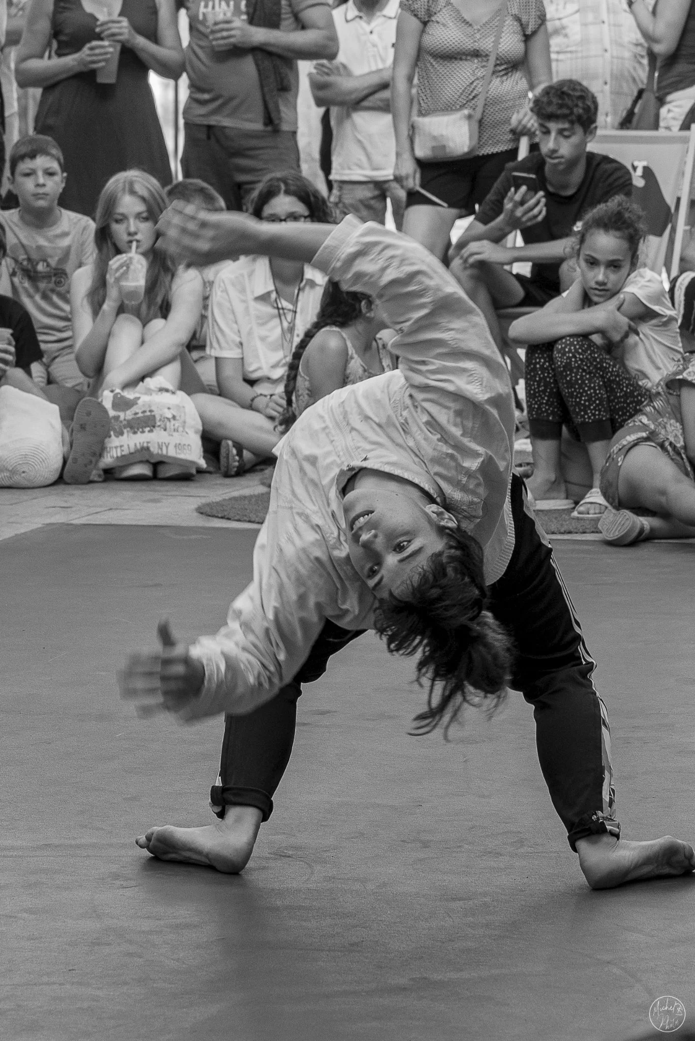 Un garçon réalise une figure de breakdance au sol, en position inversée, dans une salle remplie de spectateurs assis ou debout, certains utilisant leurs téléphones ou buvant un smoothie. La photo est en noir et blanc.