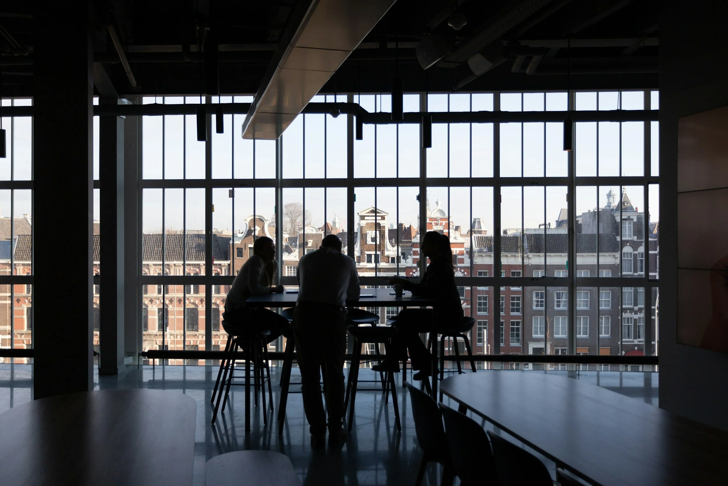 Interior of a modern office or café with large floor-to-ceiling windows showing a cityscape with historic buildings; silhouettes of three people sitting around a table near the windows.