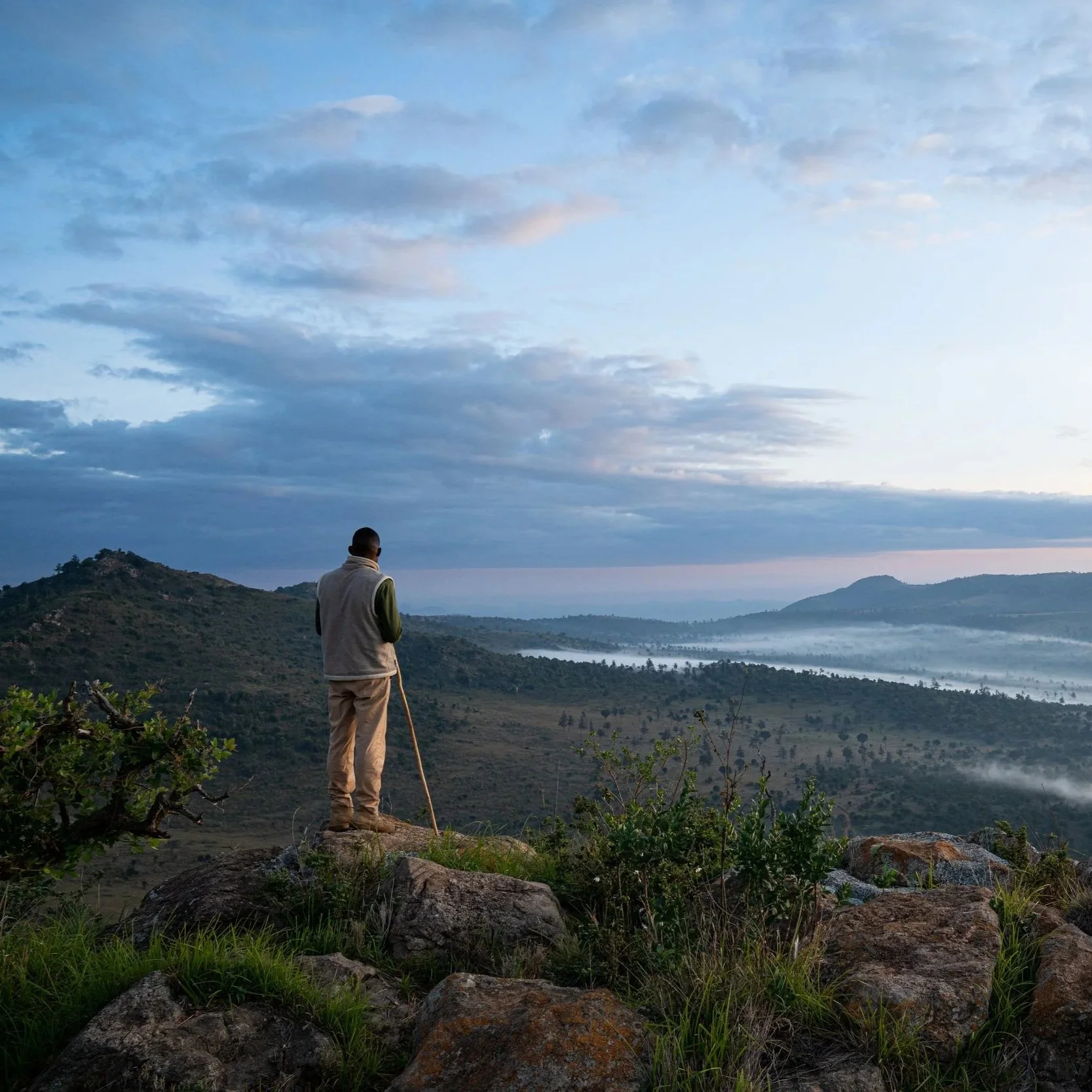 A man with a walking stick stands on a rocky hill overlooking a misty valley at dawn, with cloud-covered mountains in the distance and a sky with scattered clouds.