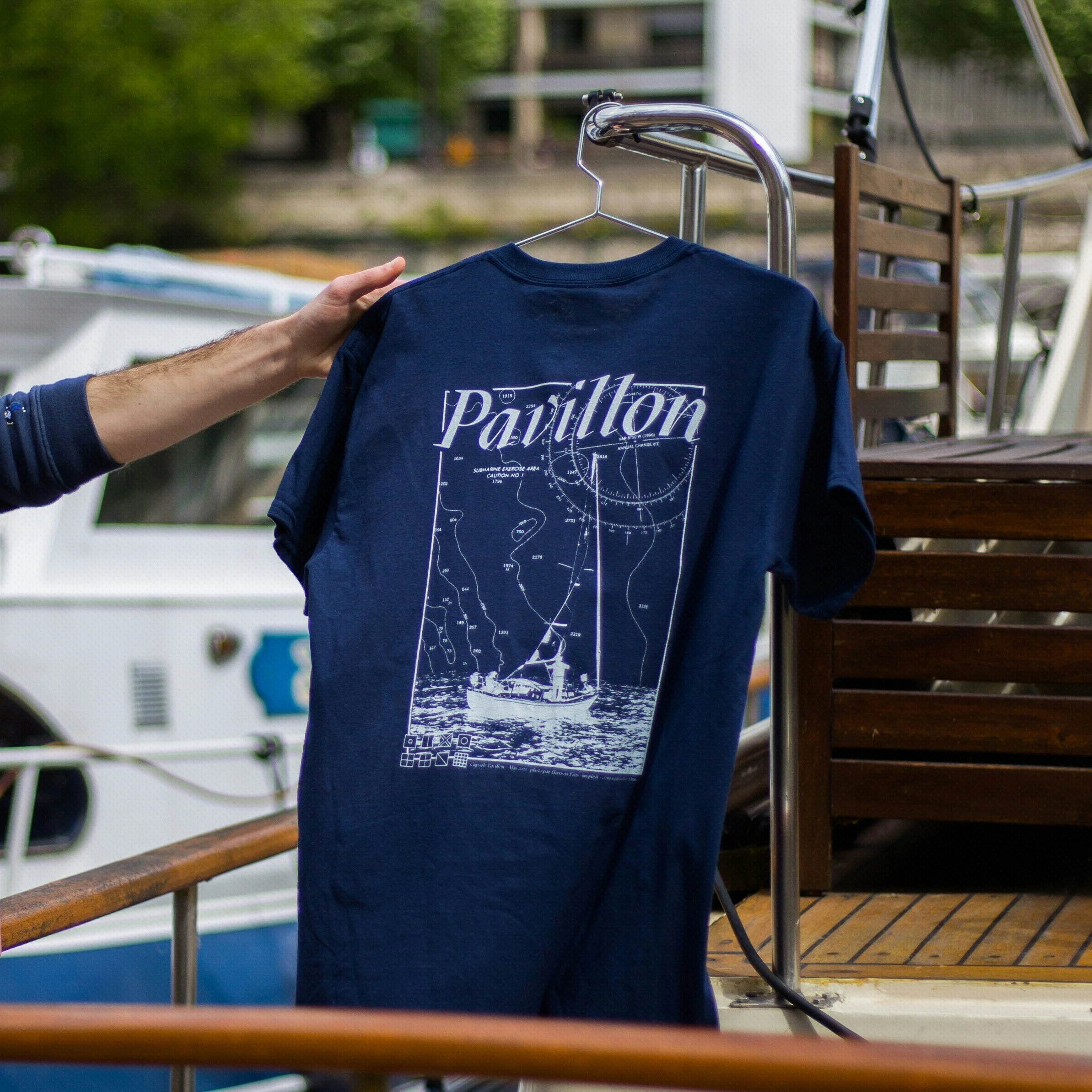 A navy blue T-shirt with white nautical diagram and text hangs on a hanger attached to a metal railing outdoors. The diagram features a submarine exercise area and a ship at the bottom. A person's hand holds the hanger.
