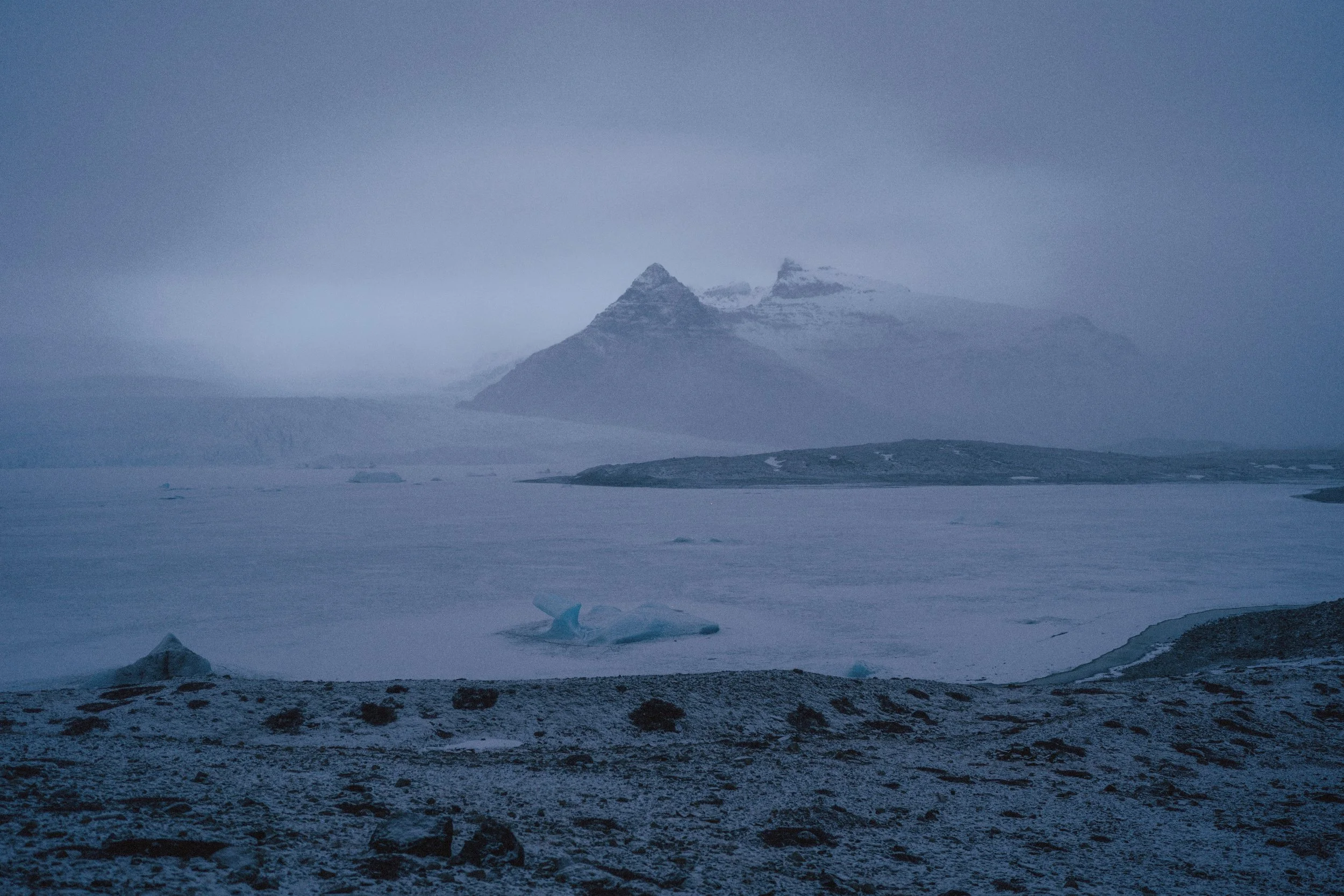 A cold, icy landscape showing a frozen body of water with icebergs in the foreground, mountainous terrain in the background, and snow-covered peaks partially shrouded in fog or low clouds.