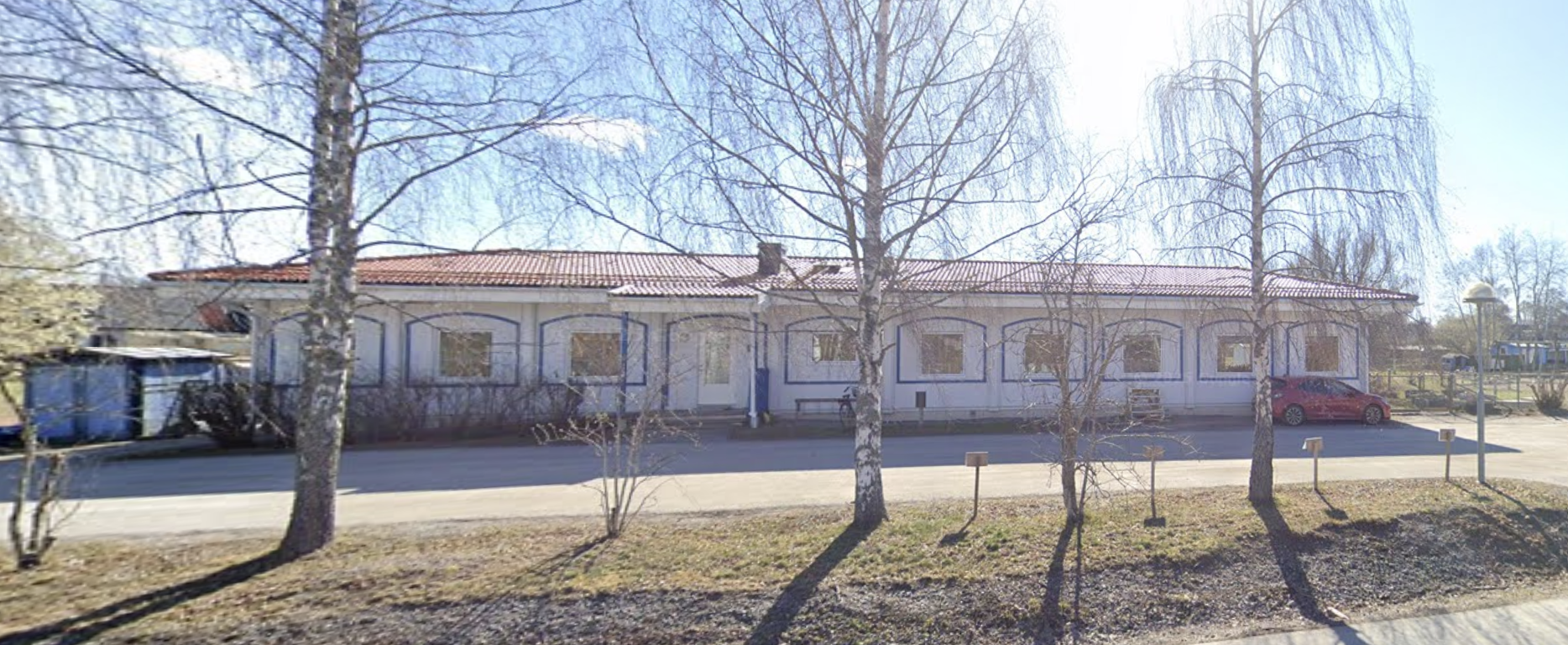 En vit byggnad med blå fönsterkarmar och ett rött tak, omgiven av några träd i vårens skugga, parkeringsplats framför och en belysningsstolpe till höger.