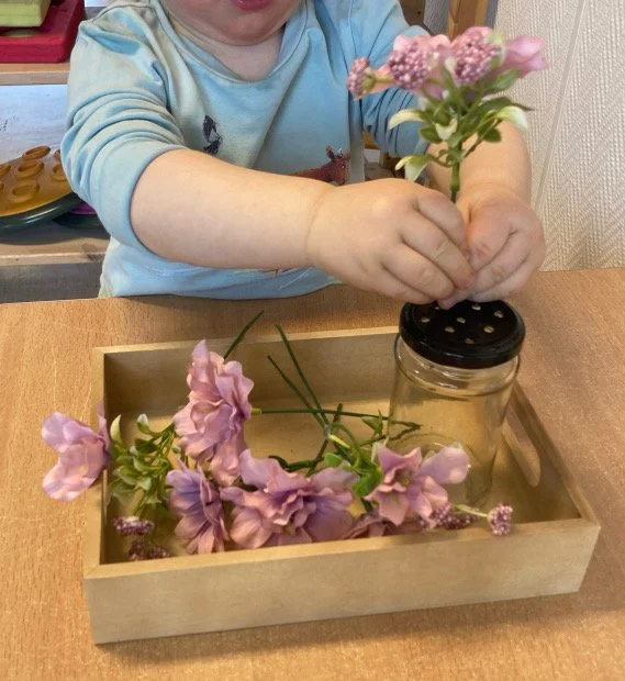 Barn som planter rosa blommor i en glasburk på ett träbord.