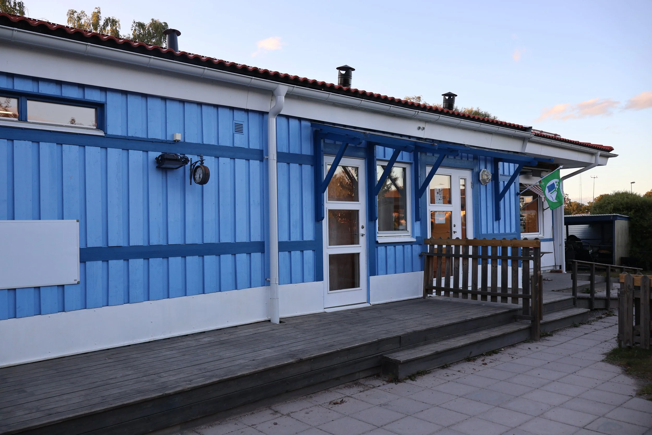 En blå byggnad med vita fönster och dörr, ett utomhusområde med trätrall och ett grönvitt flagga.