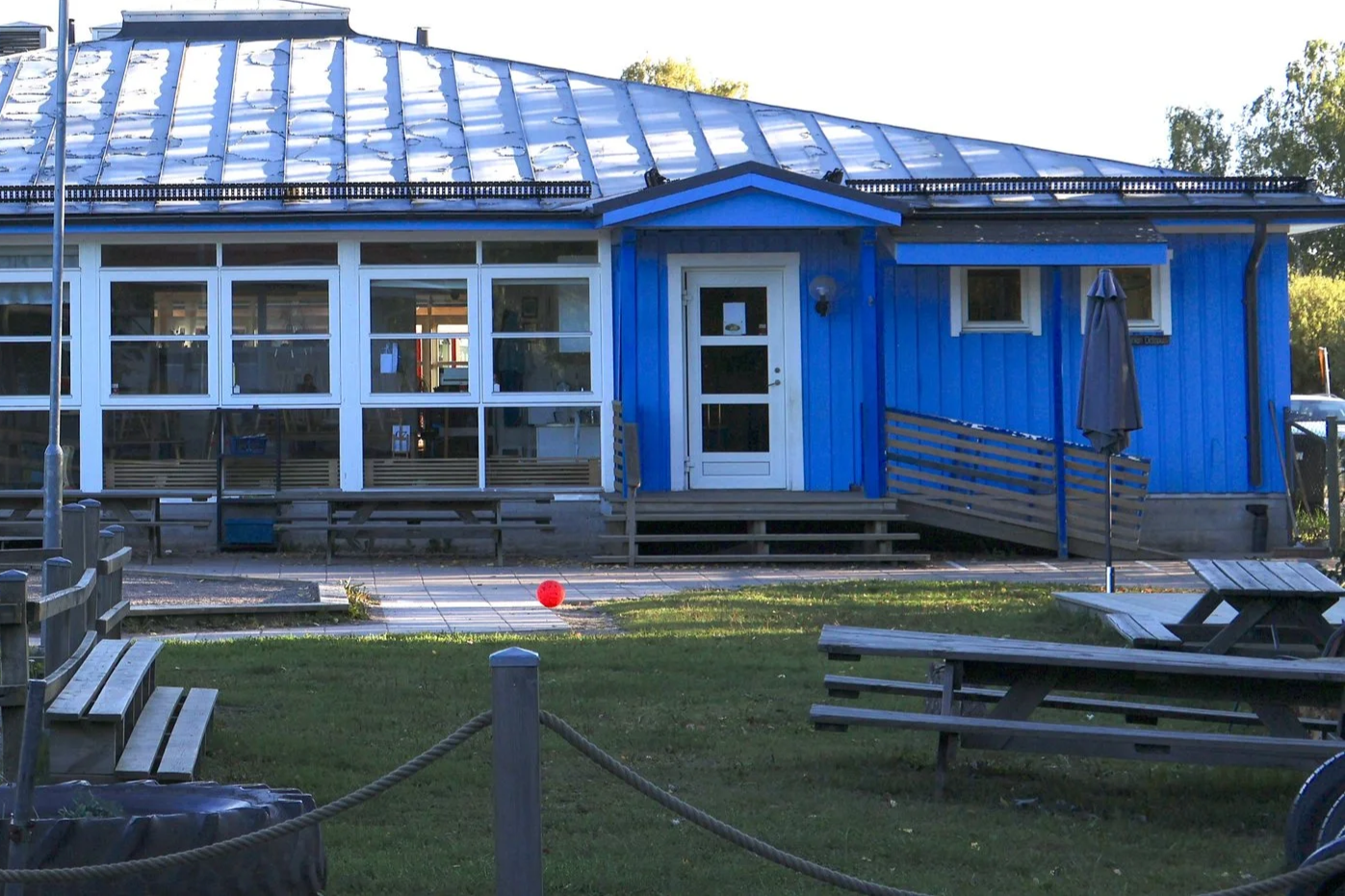 En blå trähusbyggnad med stora fönster och en vit dörr. Fronten har ett trädäck med små steg och en liten ramp, samt ett utomhusmöblemang av trä. Det finns en röd boll på marken framför huset och ett stort parasoll. Runt huset finns ett grönområde och en skyddande repstaket.