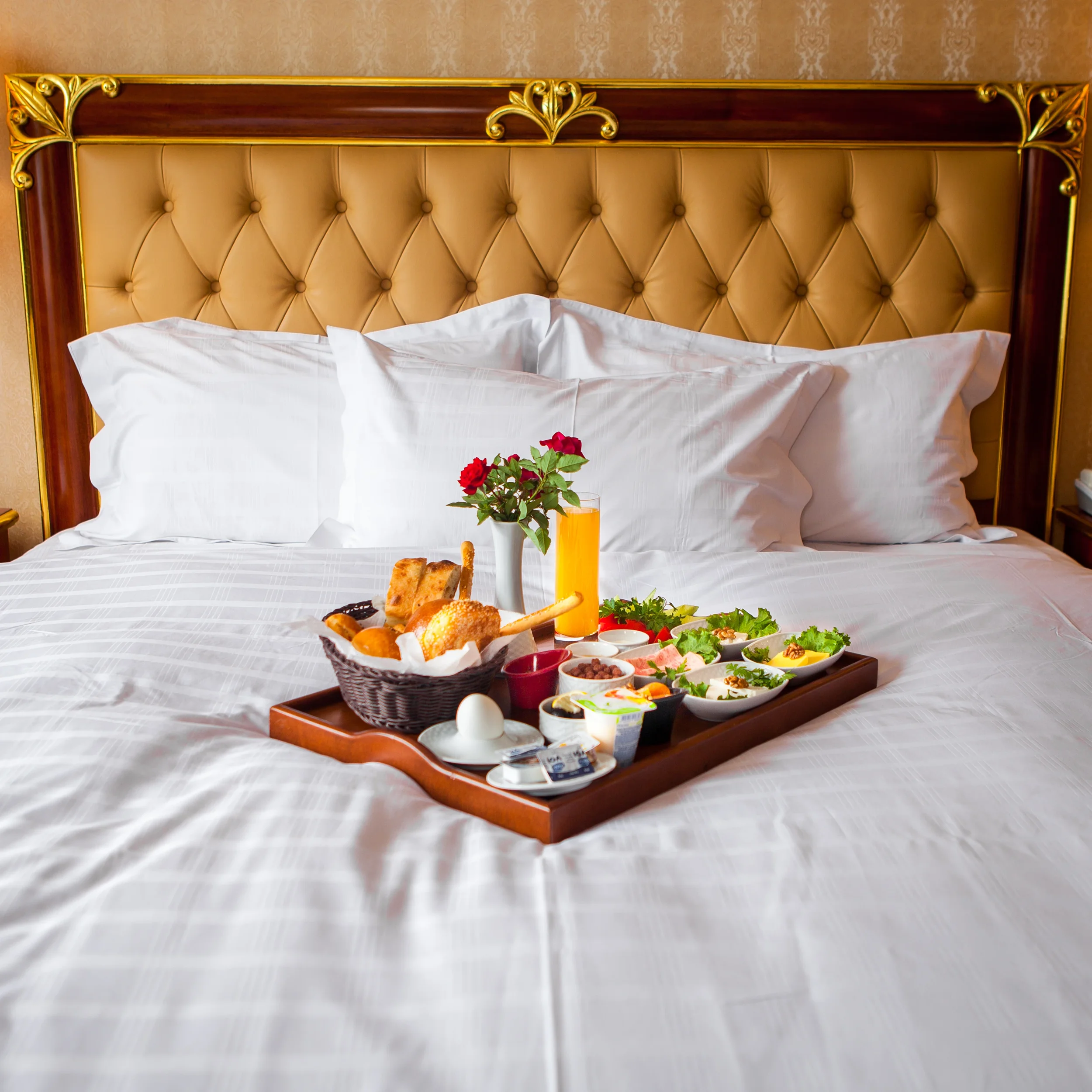 A hotel bed with a tufted, upholstered headboard featuring gold accents. On the bed, there is a tray with breakfast foods including bread, eggs, fruit, and a glass of orange juice, with a small vase of red roses.