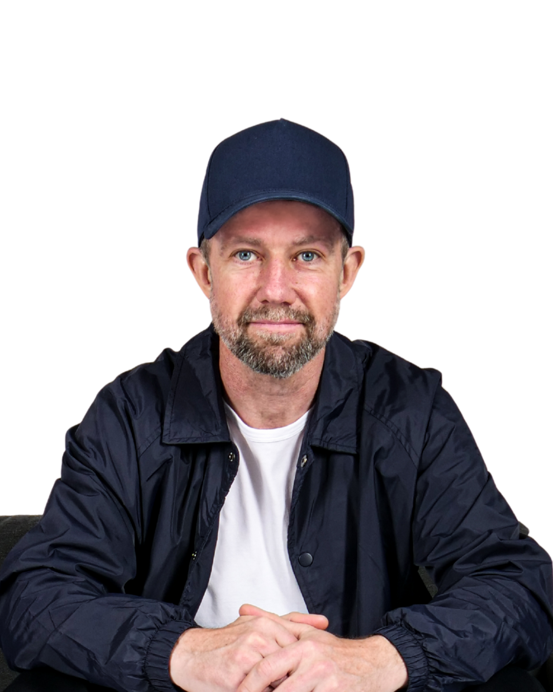 Portrait of a man with a beard and blue eyes, wearing a navy baseball cap, white t-shirt, and black jacket, sitting with hands clasped against a plain white background.