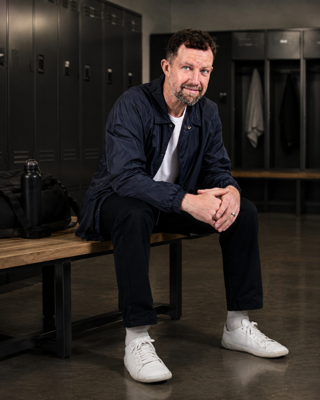 Andrew Sillitoe sitting on a wooden bench in a locker room, wearing a dark jacket, white t-shirt, black pants, white sneakers, and gray socks. There are black lockers behind him and a black water bottle and bag on the bench.