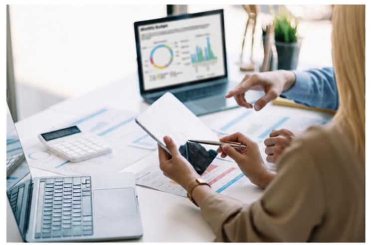 Two women are working at a desk cluttered with papers, a calculator, a laptop, and a tablet. One woman is pointing at the tablet, while the other is holding the tablet and using a stylus. A laptop screen in the background displays graphs and charts.