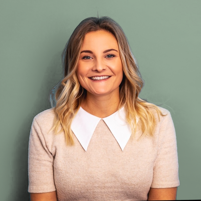 A young woman with shoulder-length blonde hair smiling, wearing a beige top with a white collar, standing against a green background.
