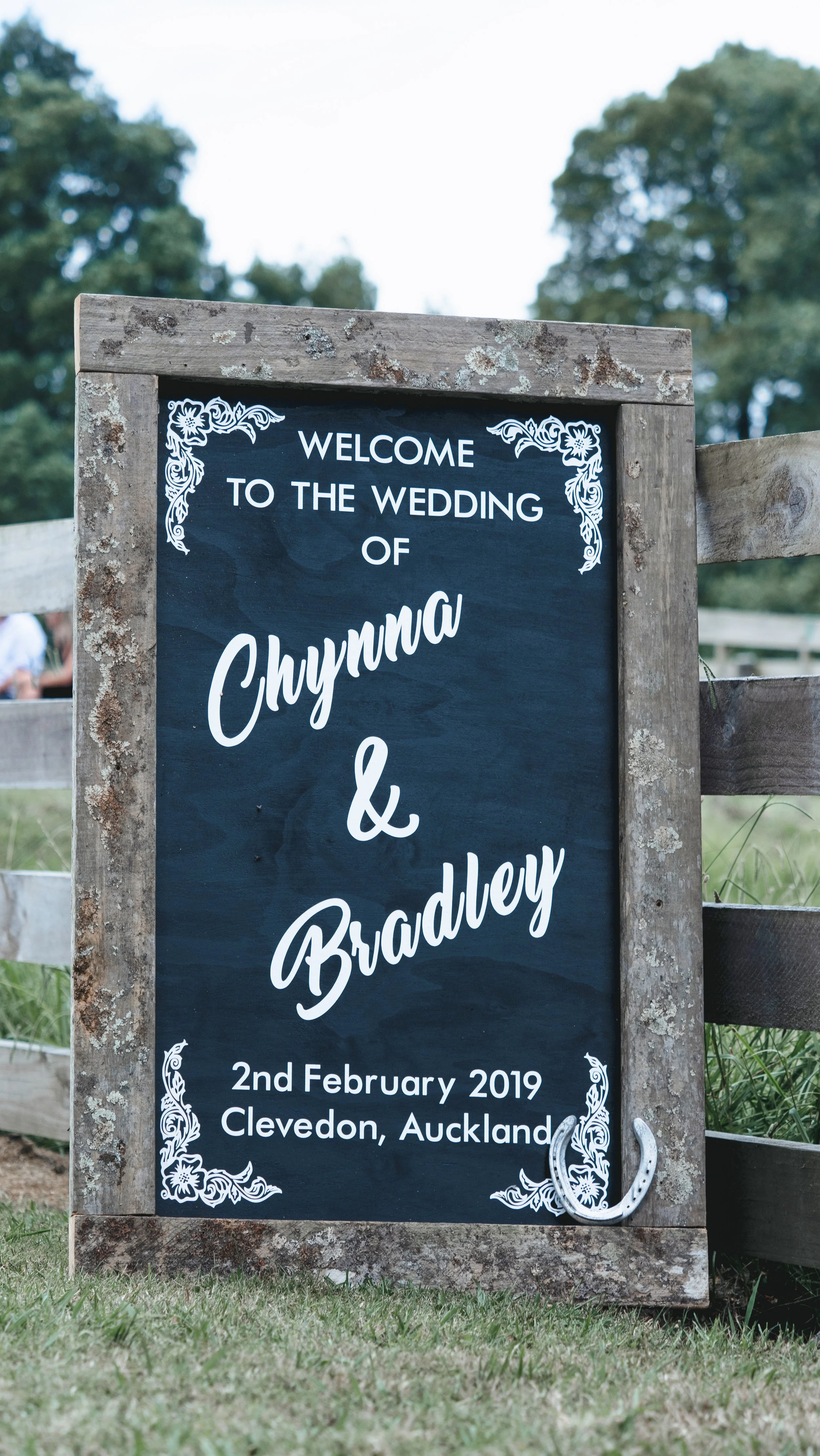 A chalkboard-style sign with a wooden frame at an outdoor wedding event. The sign reads, 'Welcome to the wedding of Chynna & Bradley, 2nd February 2019, Clevedon, Auckland,' with decorative floral designs in the corners.