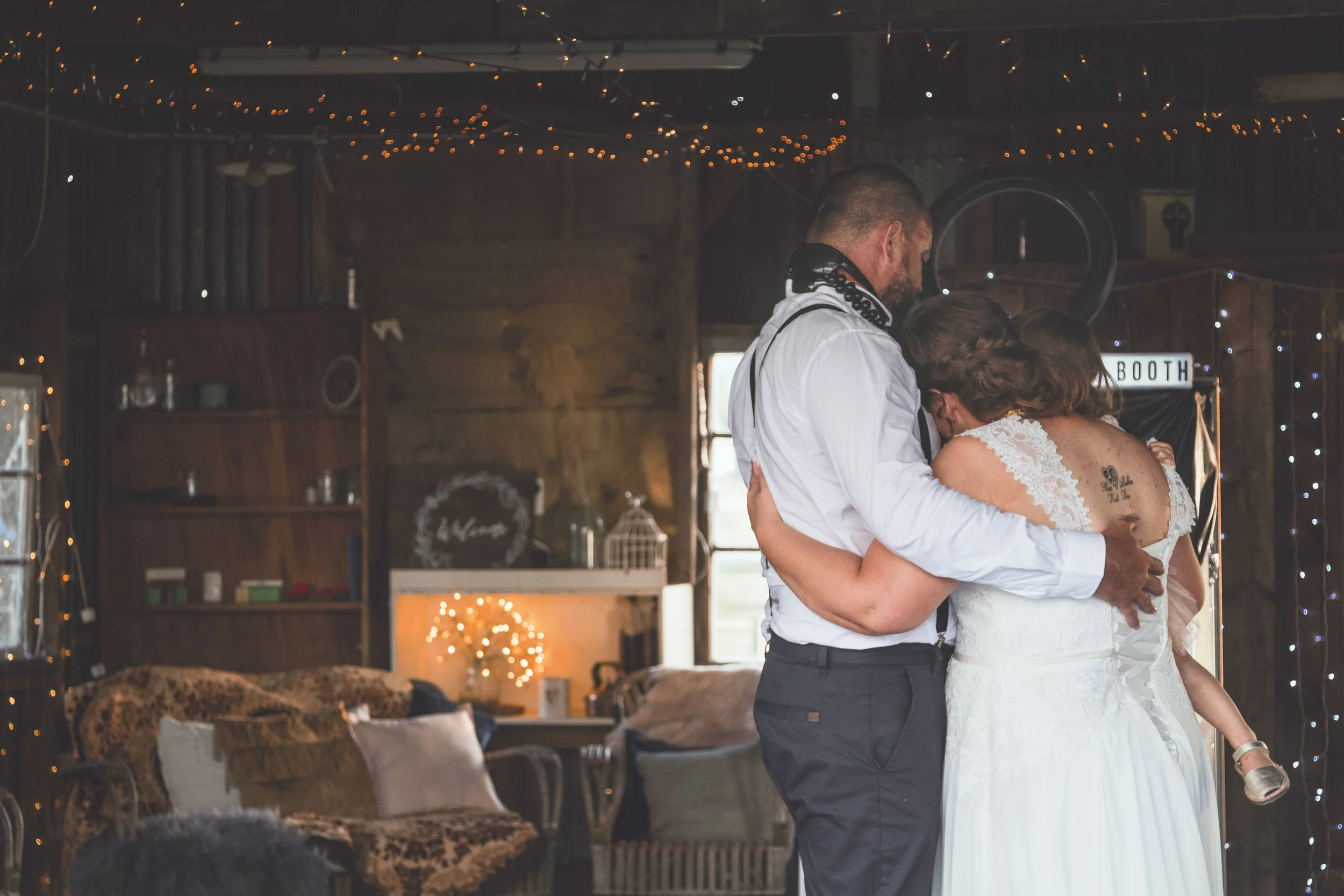 A couple dancing closely at a wedding reception in a rustic venue decorated with string lights and warm lighting.