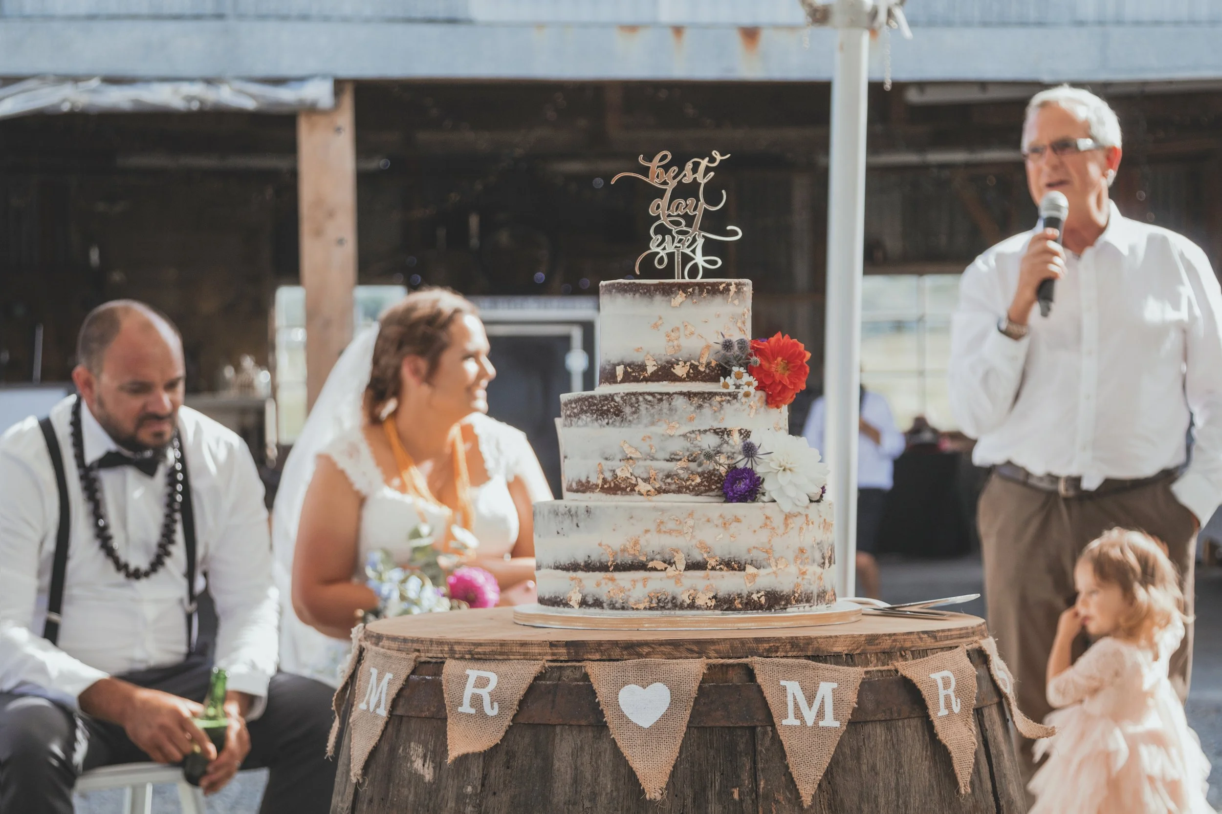Wedding reception with a rustic theme, featuring a tall, semi-naked wedding cake decorated with flowers and a topper that says 'Last Day Ever'. Guests are seated at tables, including a woman in a white dress, a man in a white shirt, and another man i