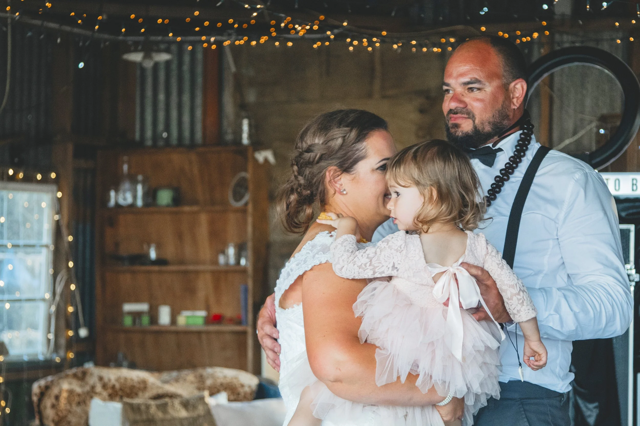 A woman in a wedding dress holding a young girl in a white dress with a bow, as a man in a white shirt and suspenders looks on. They are indoors with string lights on the ceiling and wooden walls.