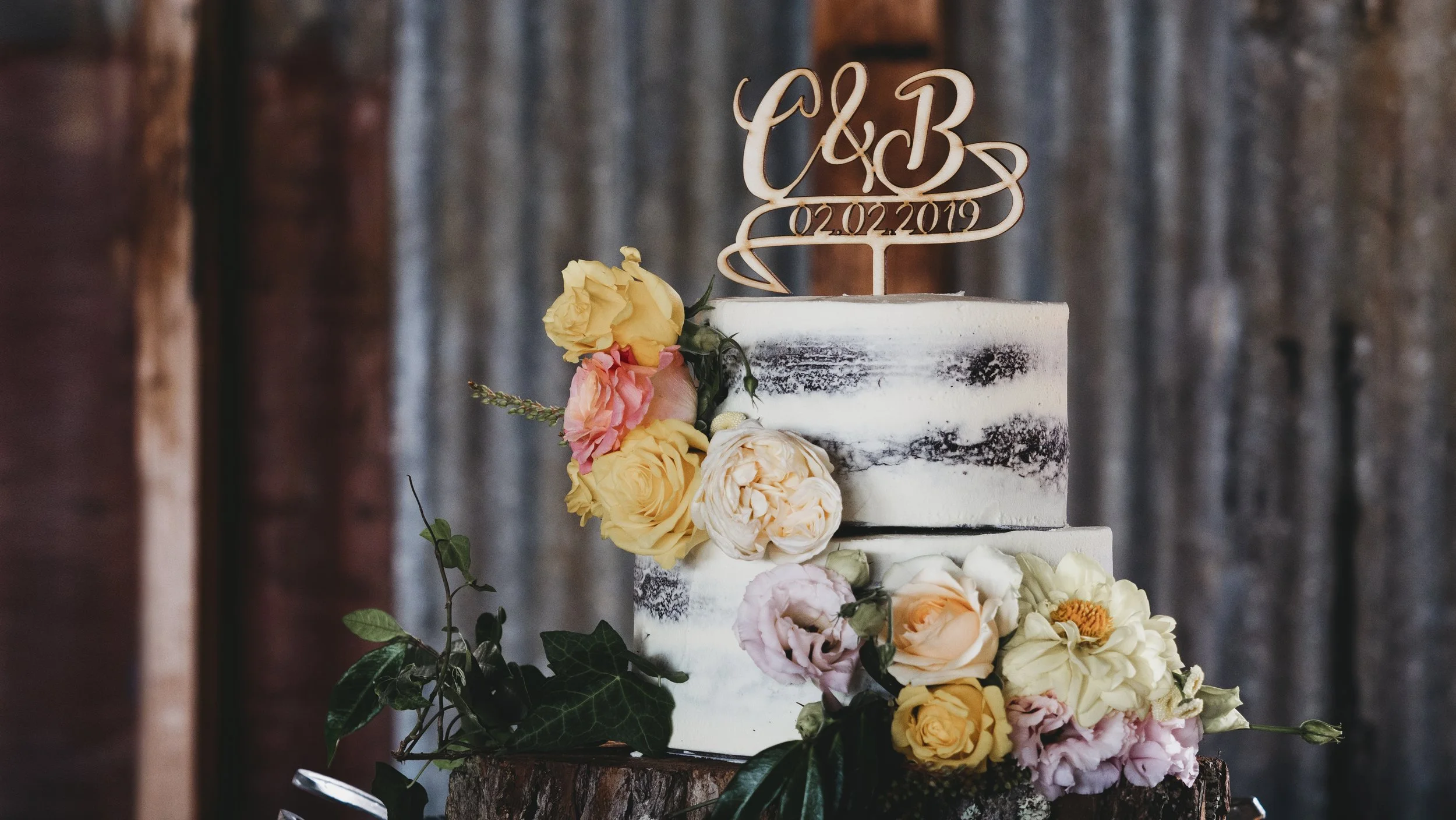 Wedding cake decorated with fresh yellow, pink, and cream flowers, with a cake topper featuring initials and a date.
