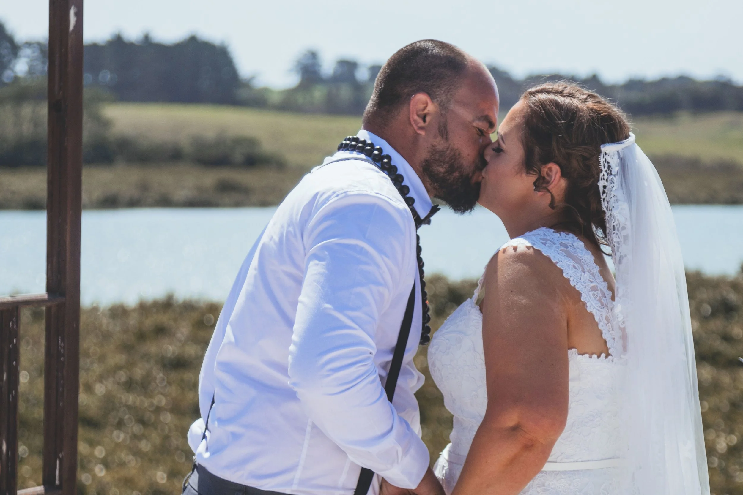 A man and a woman are kissing outdoors, dressed in wedding attire, near a body of water with a scenic background of trees and open landscape.