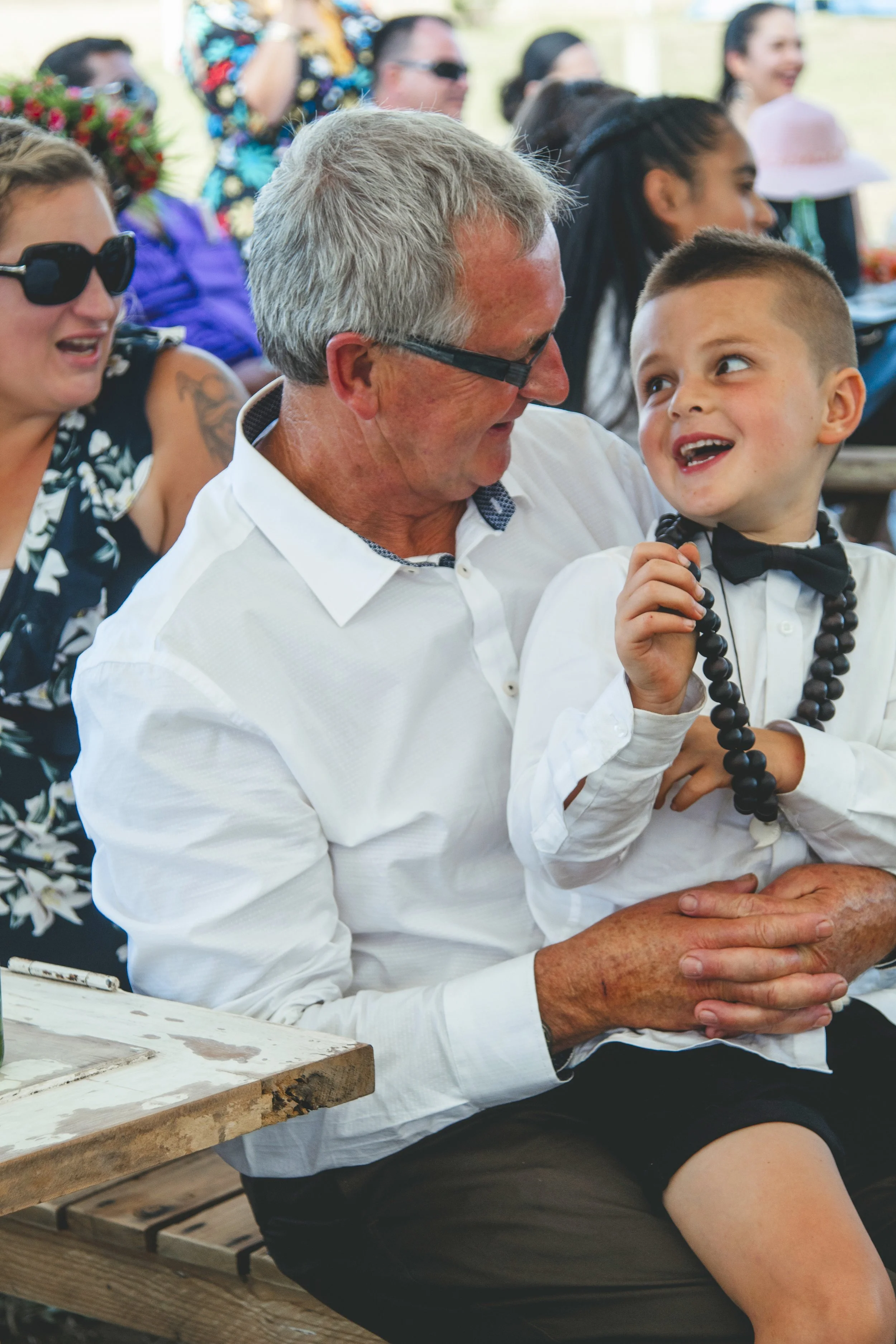 A group of people gathered at an outdoor event, smiling and engaging with each other. A man with gray hair and glasses is seated next to a young boy wearing a white shirt and black bow tie, both smiling and interacting. In the background, several wom