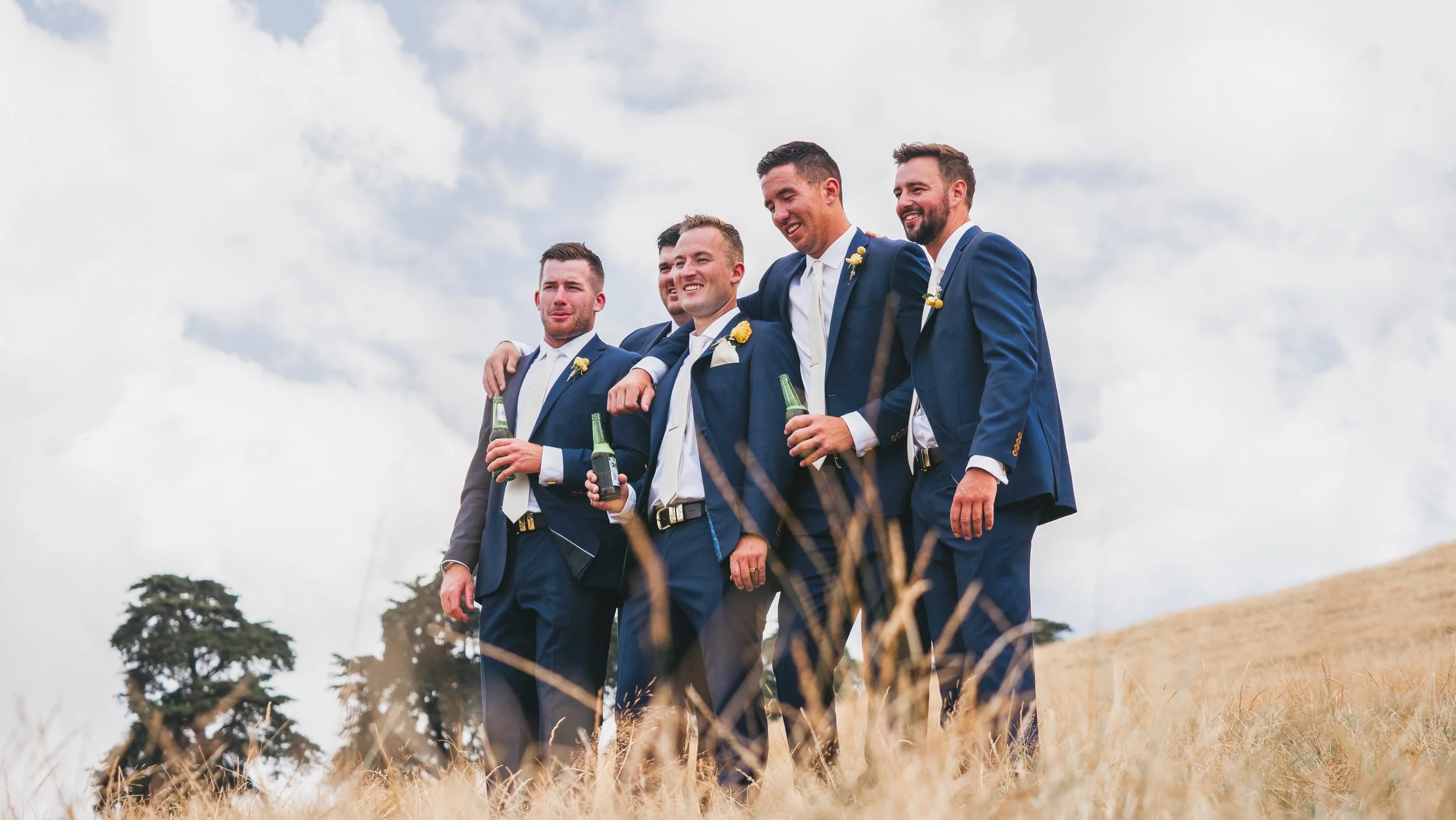 A group of five men dressed in blue suits and white shirts, standing outdoors on a cloudy day, smiling and holding beer bottles, with one arm around each other's shoulders.