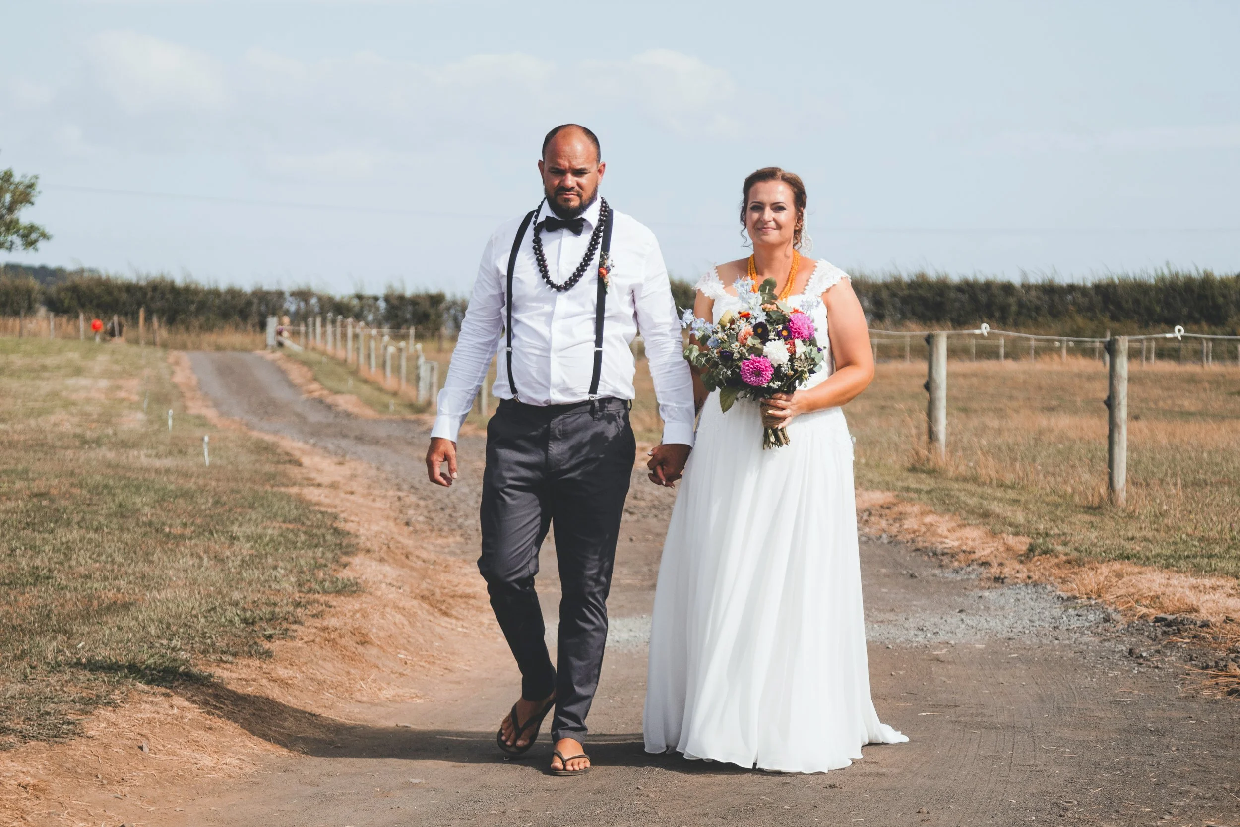 A bride and groom walking hand in hand outdoors on a dirt path, with a fence and grassy landscape in the background under a partly cloudy sky. The bride is holding a colorful bouquet and wearing a white wedding dress, while the groom is dressed in a 