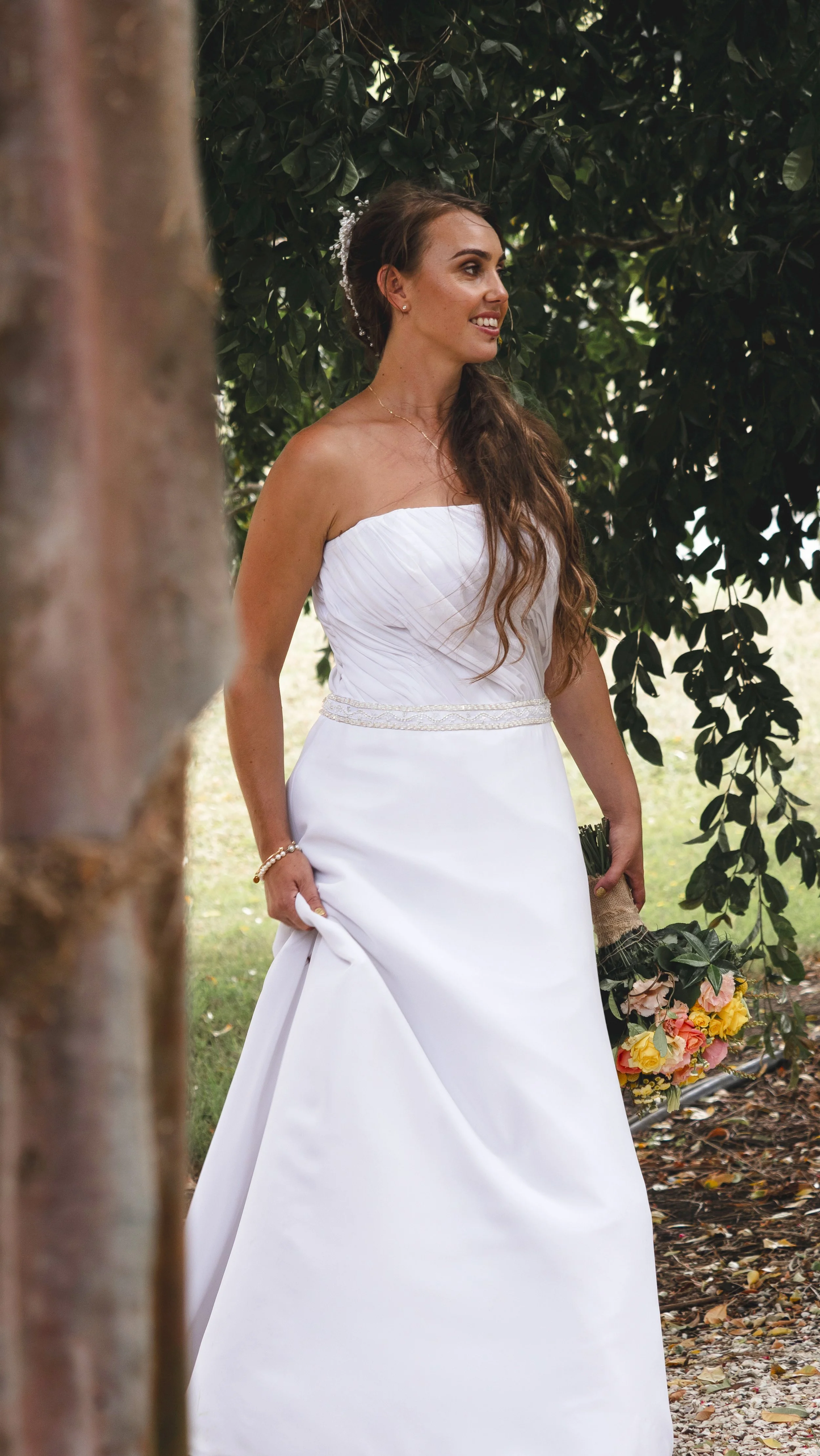 A woman in a white wedding dress standing outdoors near trees, holding a bouquet of flowers.