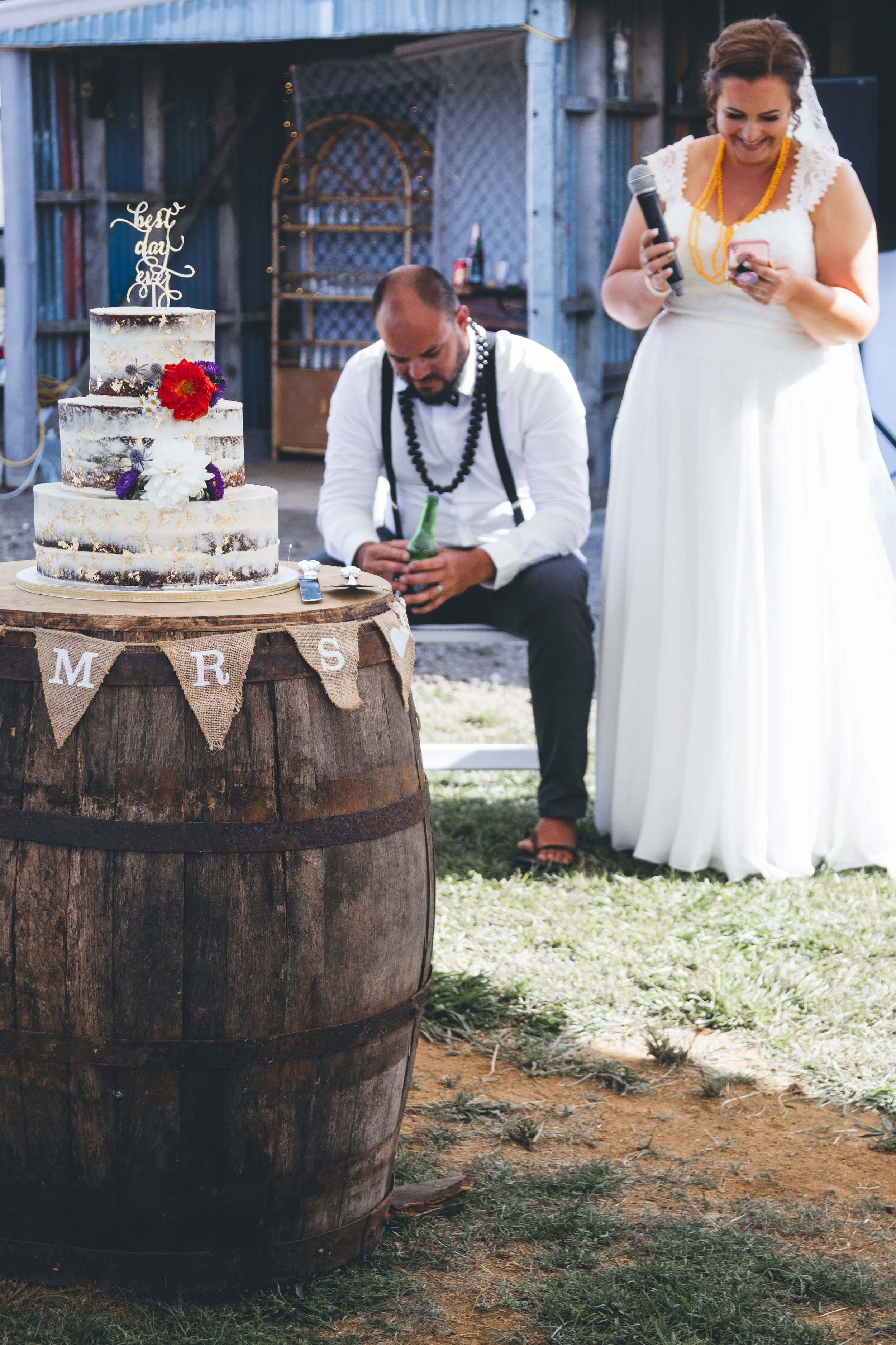 A bride in a white dress and a man in a white shirt with suspenders and a bow tie, sitting behind a rustic wooden barrel decorated with a burlap banner reading 'Mrs' and flowers, in an outdoor setting with a wooden fence and a wedding cake decorated 
