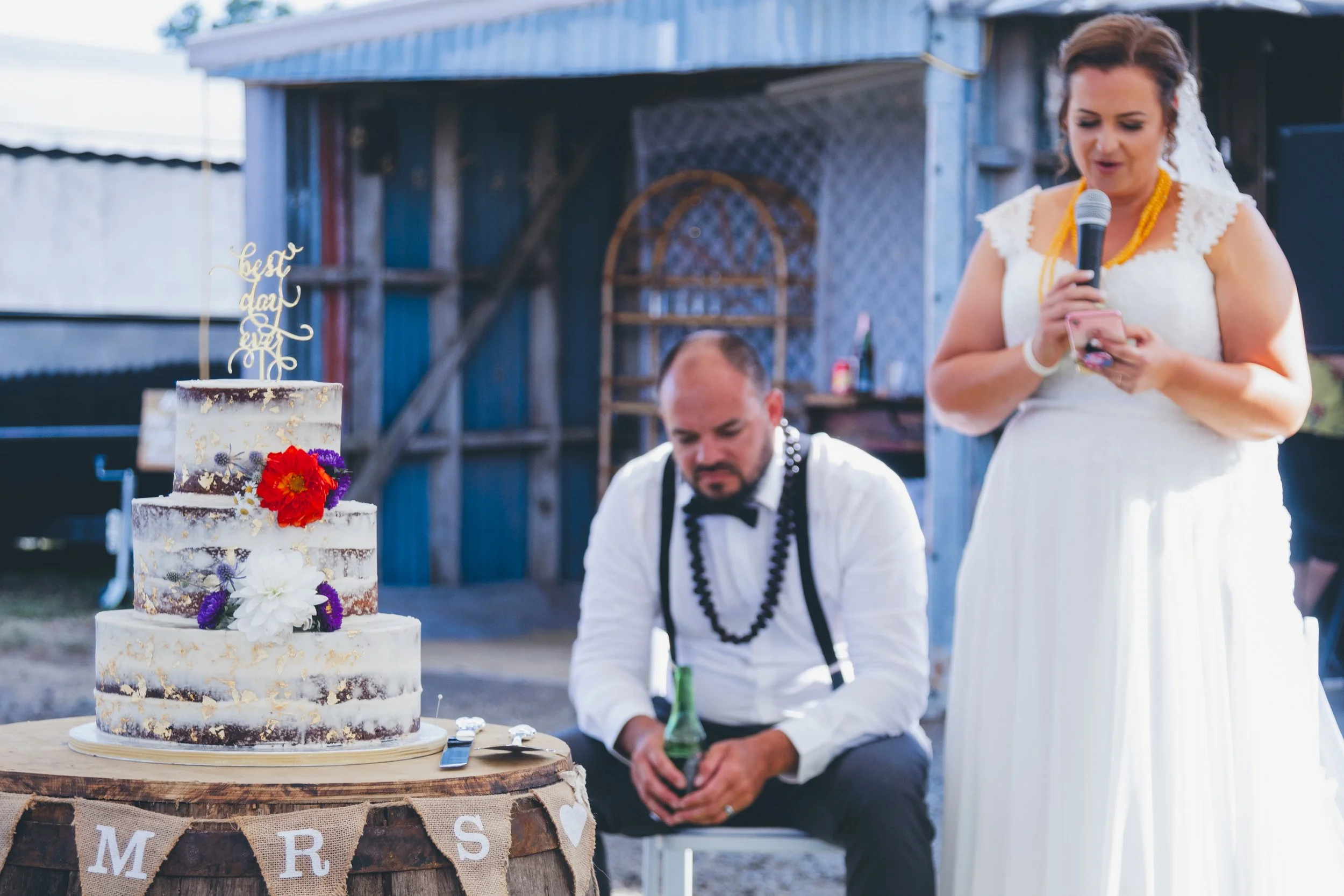 Wedding celebration with a wedding cake decorated with flowers, a woman in a white dress holding a microphone and reading, and a man sitting nearby in an outdoor setting.