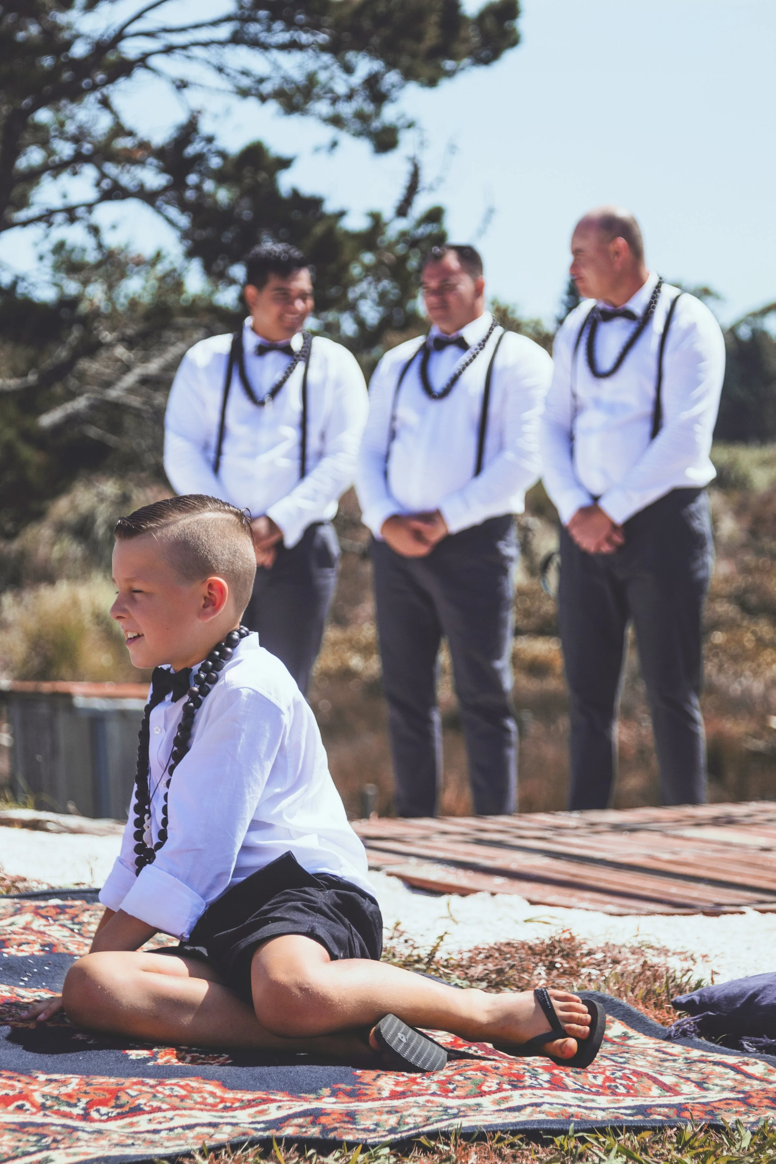 Young boy sitting cross-legged on a patterned rug outdoors, with three men dressed in white shirts, black bow ties, suspenders, and necklaces standing behind him, smiling and talking.