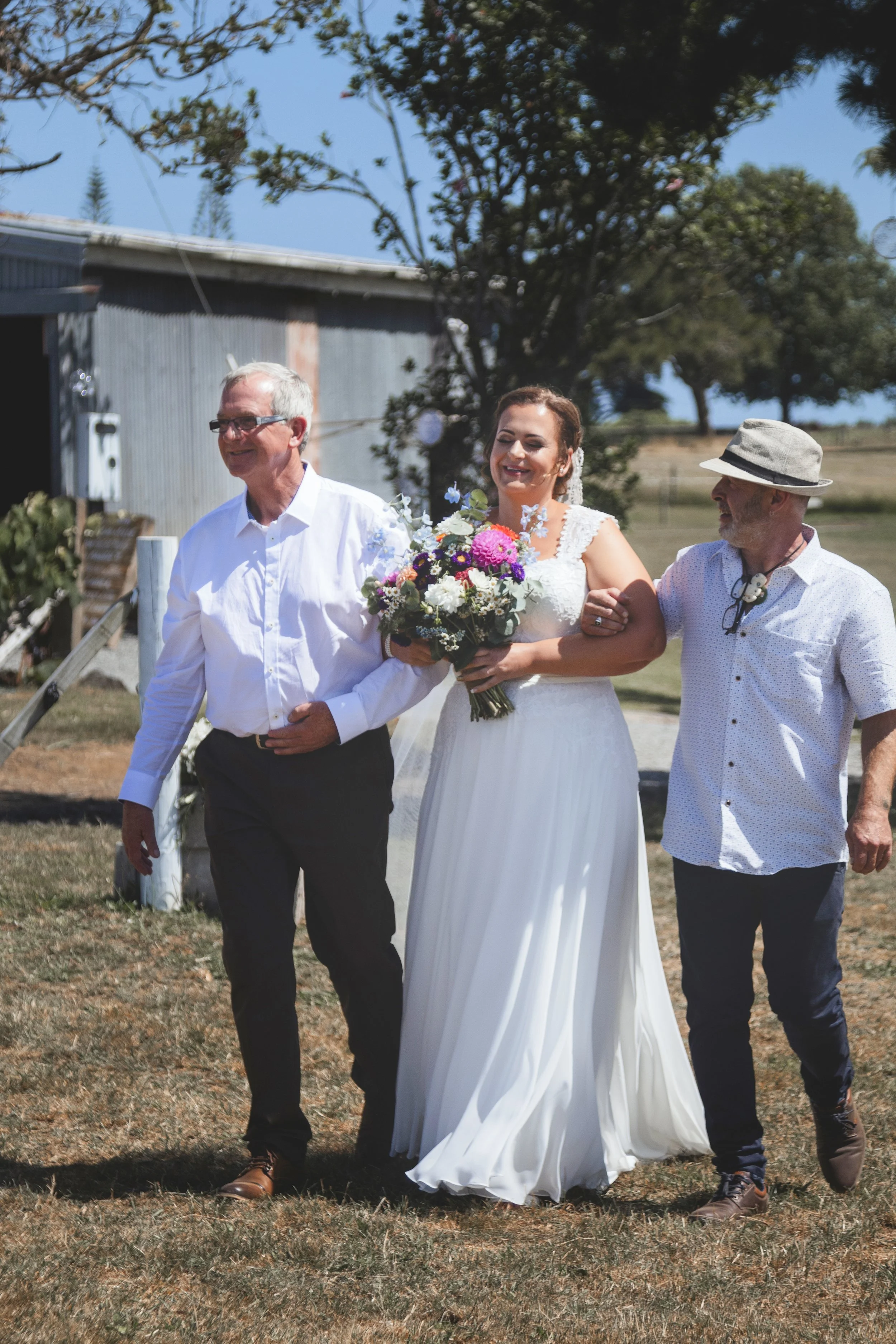 A bride in a white dress holding a bouquet of flowers walking between two older men outdoors during a sunny day.