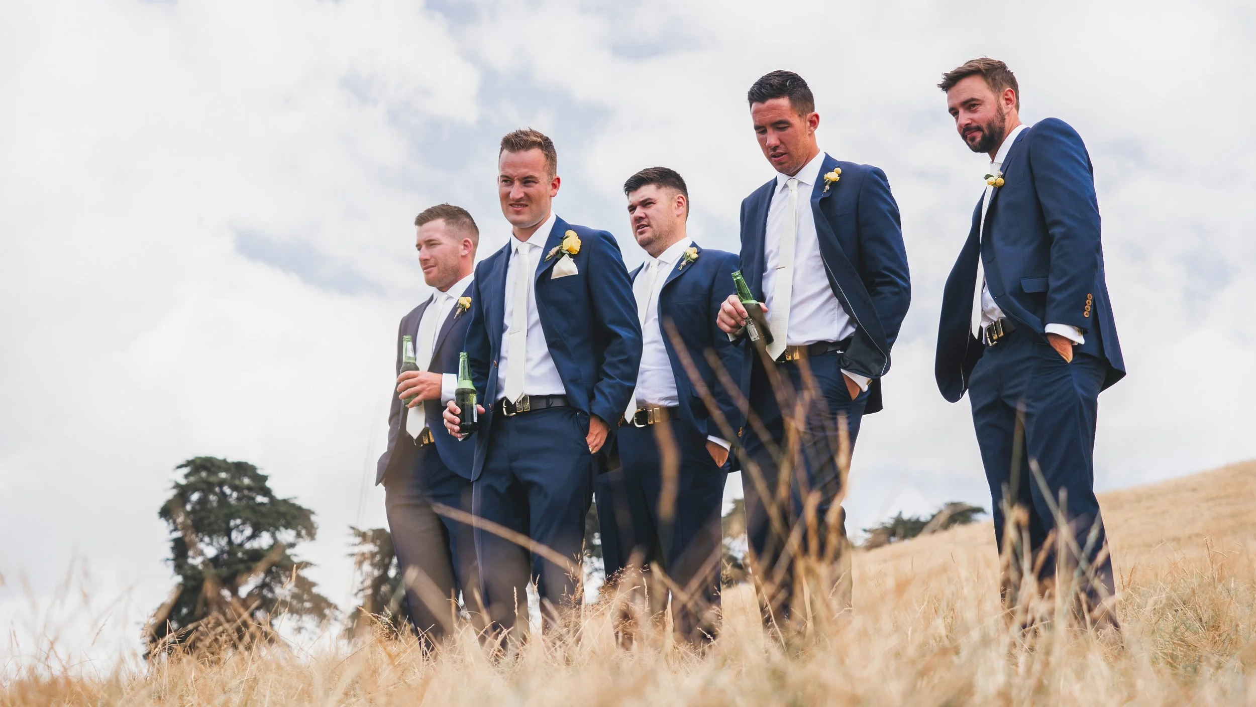 Group of six men in blue suits and white shirts, holding beer bottles, standing outdoors in a field with tall grass and trees under cloudy sky.