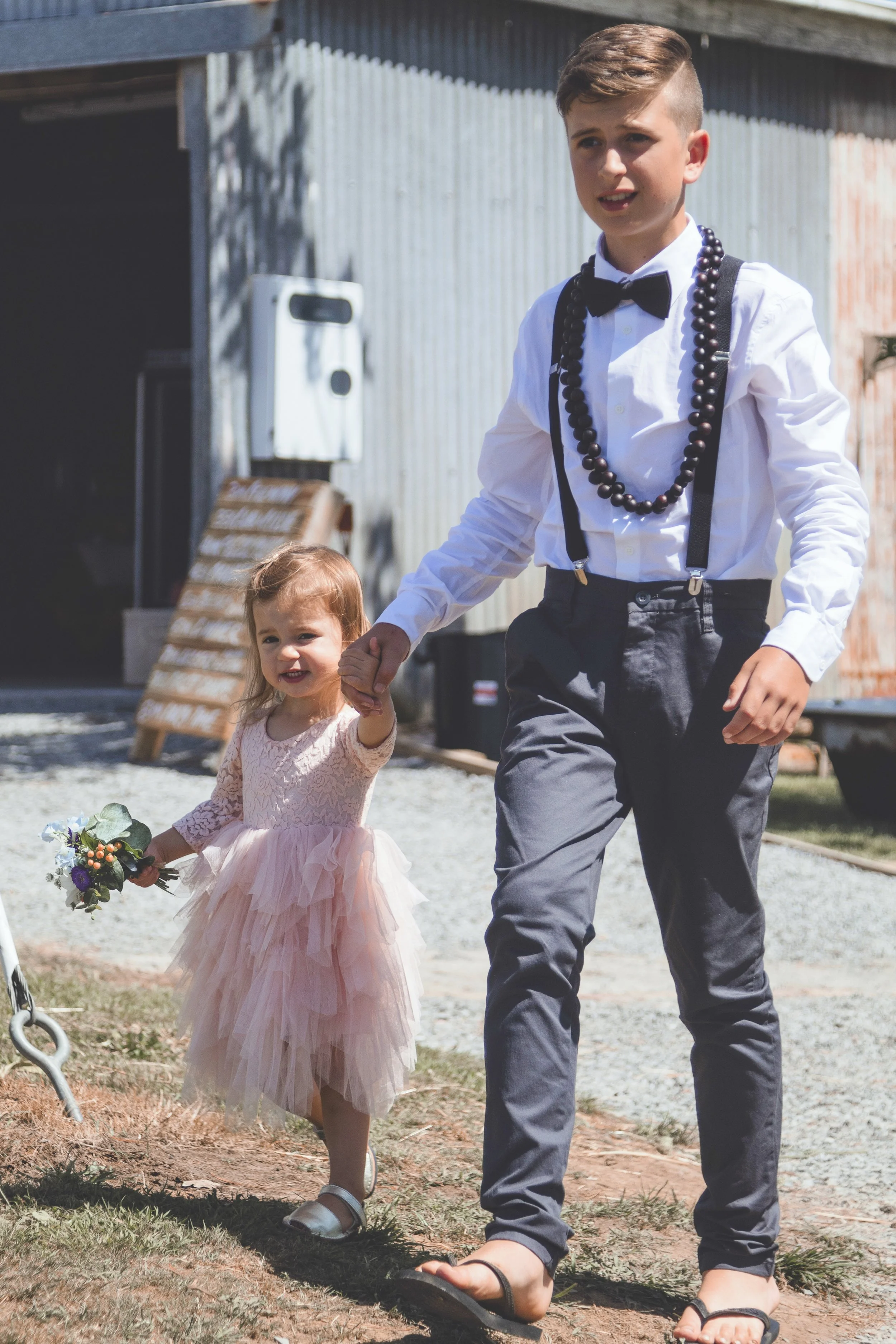A young man in a white shirt, black bow tie, suspenders, and gray pants is holding hands with a little girl. The girl is wearing a pink dress with layers of tulle, white sandals, and is holding a bouquet of flowers. They are outdoors near a barn-like