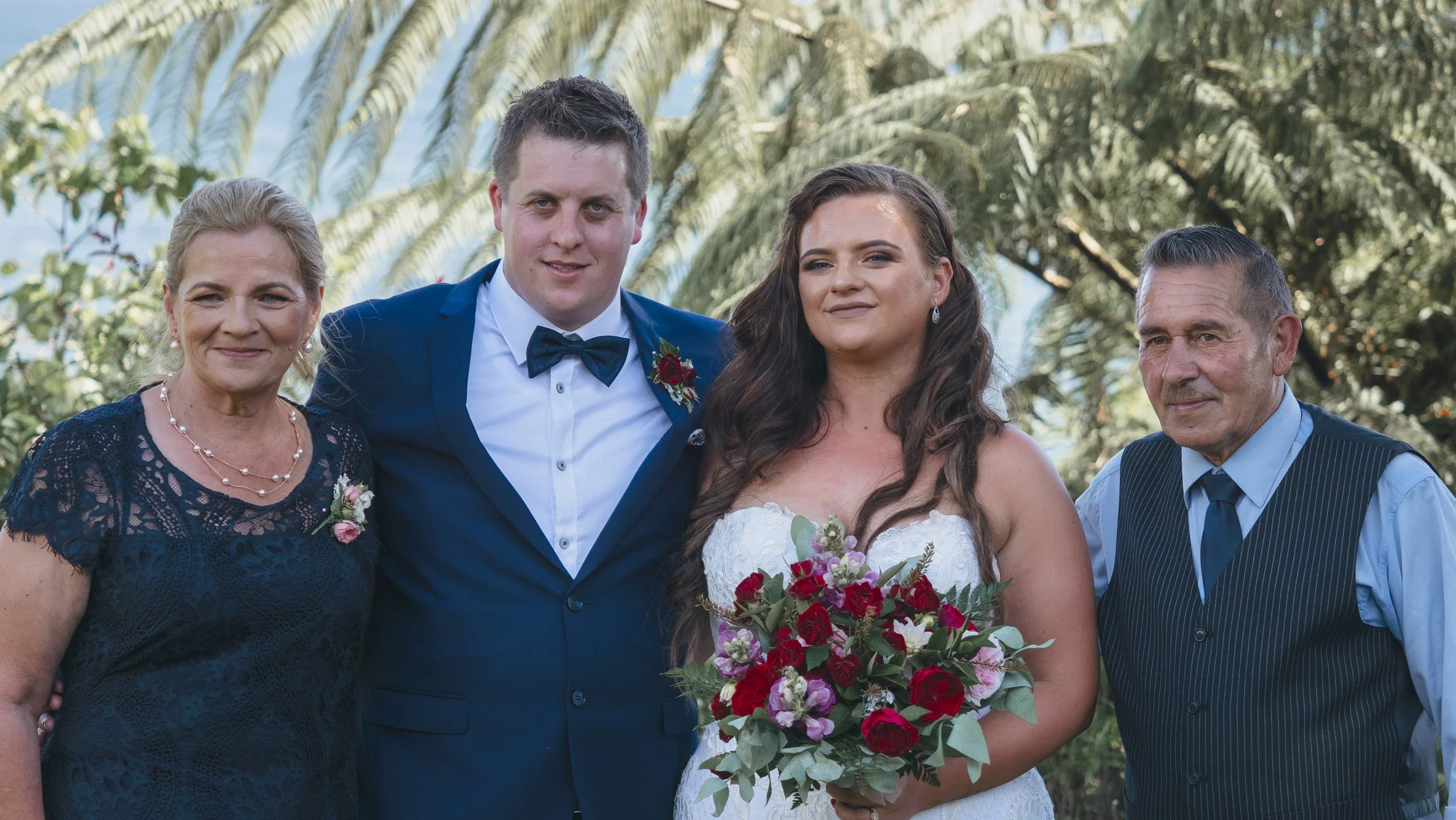 A family celebrating a wedding outdoors, with a bride holding a bouquet of colorful flowers, surrounded by two men and a woman.