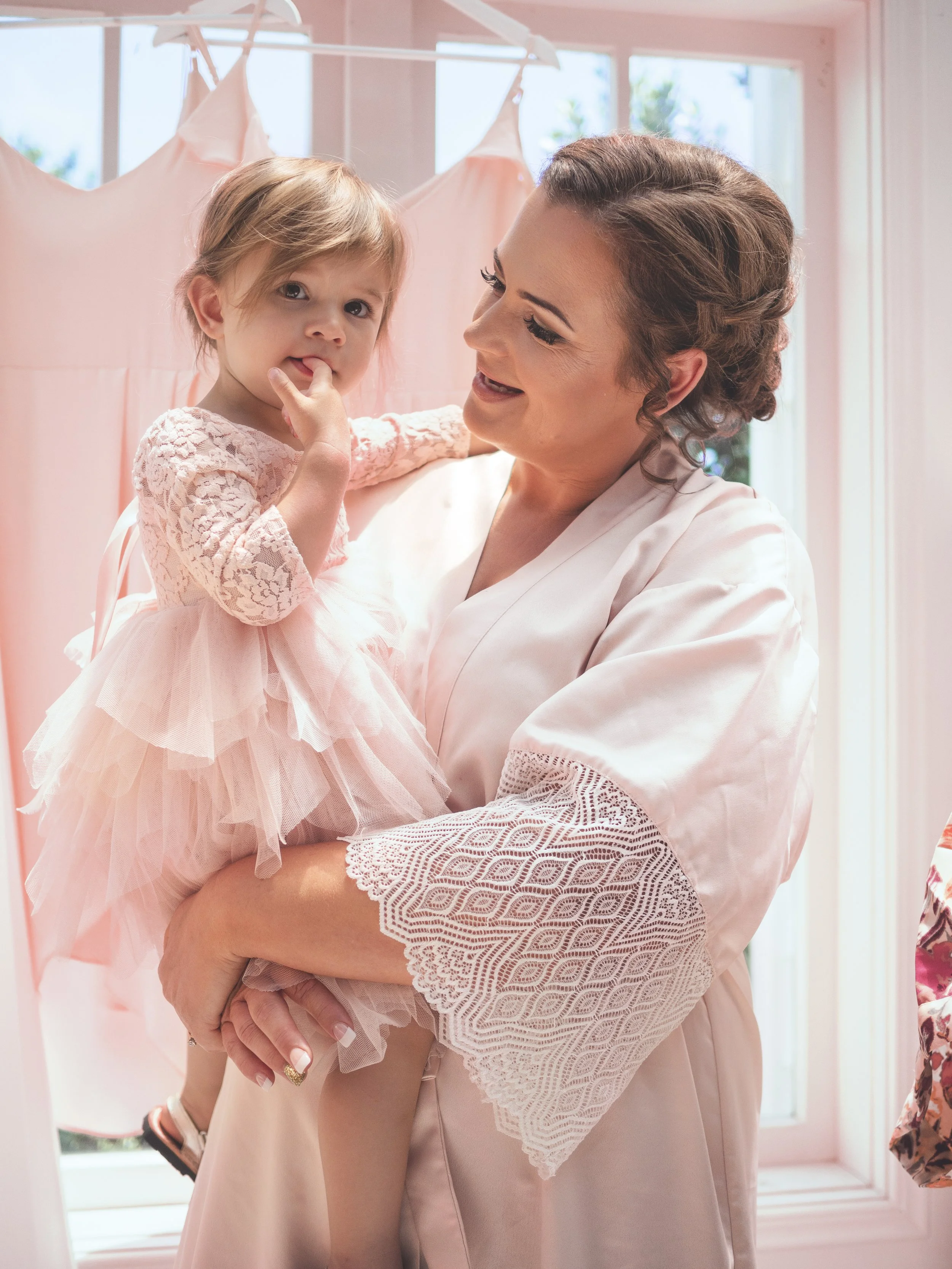 A woman holding a young girl inside a room with pink and white decor, pink dresses hanging on a rack, and a window with a view of greenery outside.