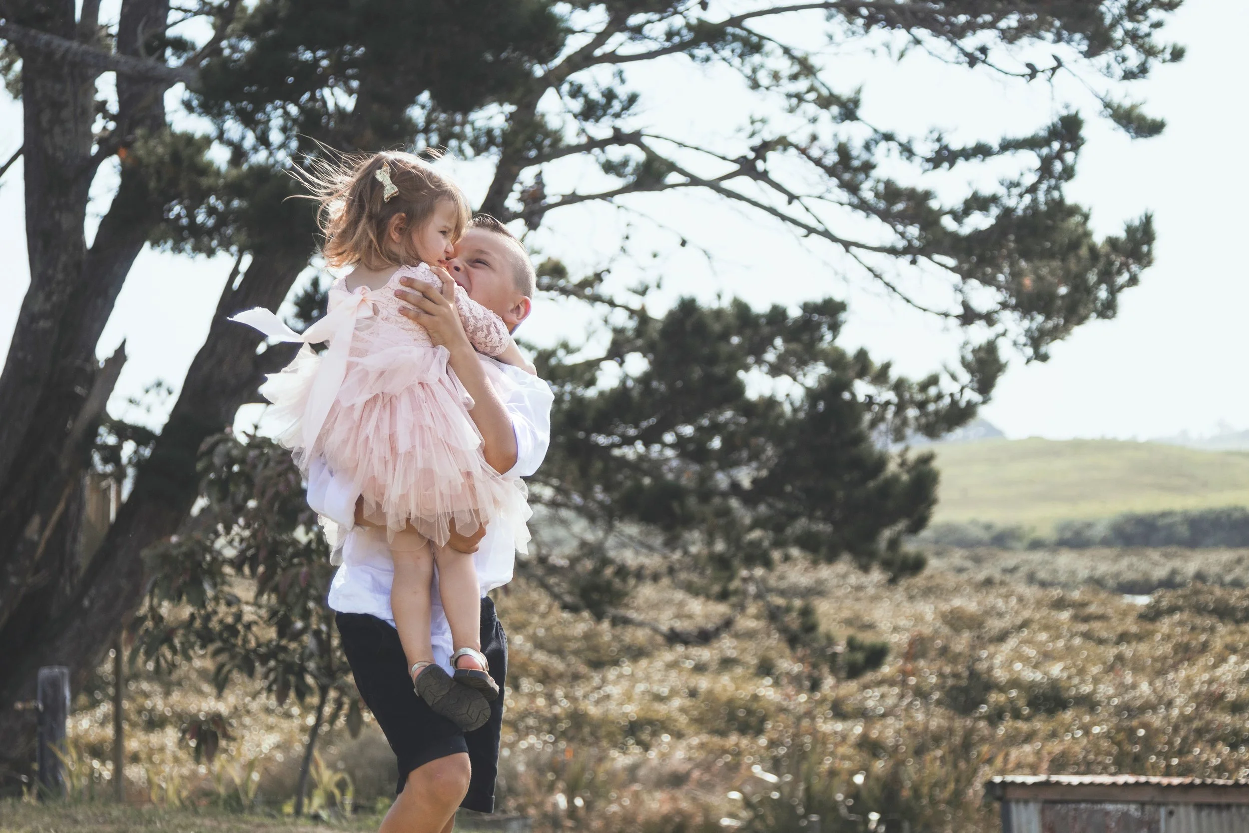 A person is standing outdoors, holding a young girl in the air. The girl is dressed in a pink dress with ruffles, and her hair is adorned with a hairclip. Trees and a grassy area are visible in the background, and the scene appears to be a park or na