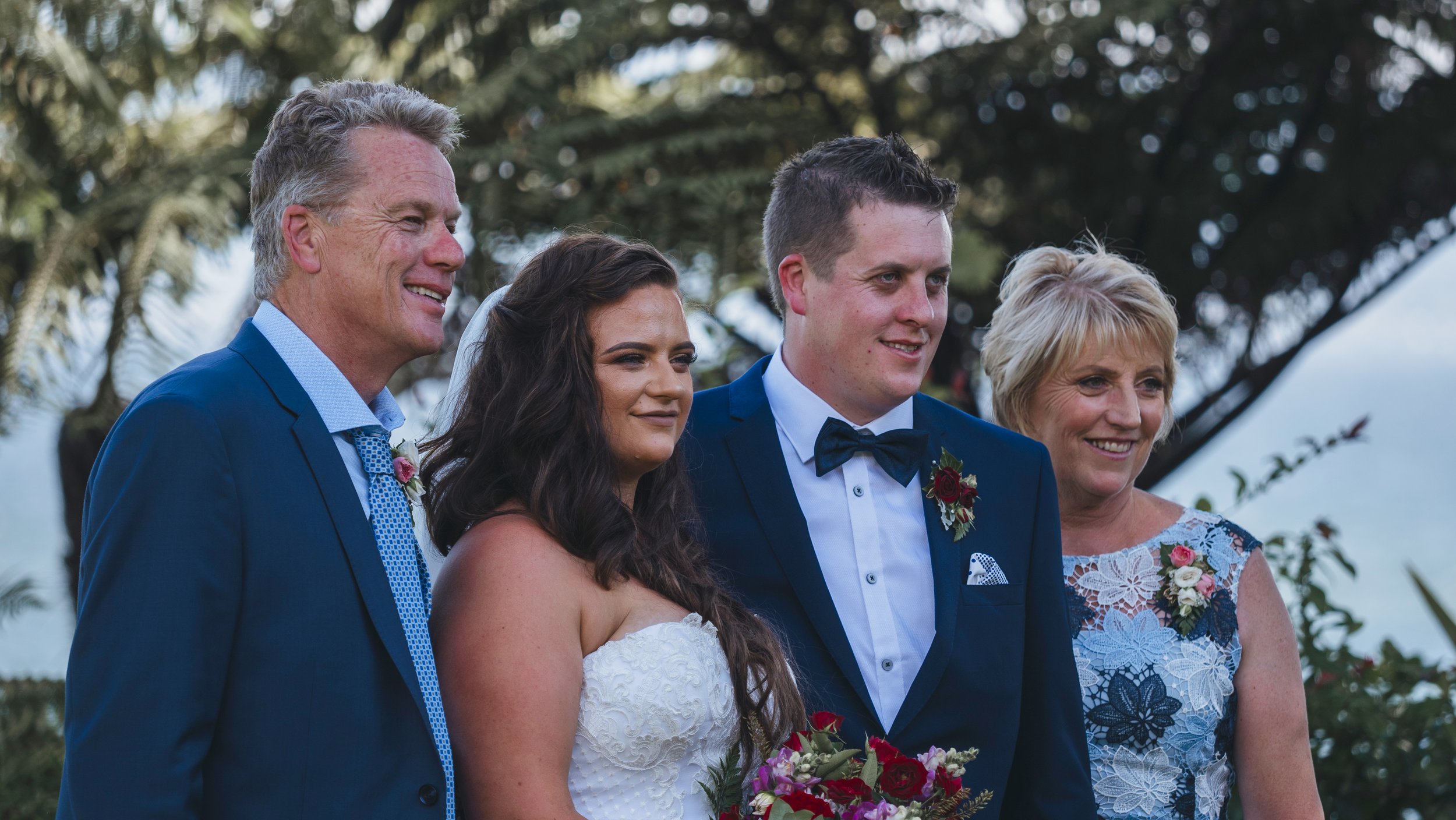 Group of five people at a wedding, including a bride in a white dress, a groom in a tuxedo, and three older adults, outdoors with trees in the background.