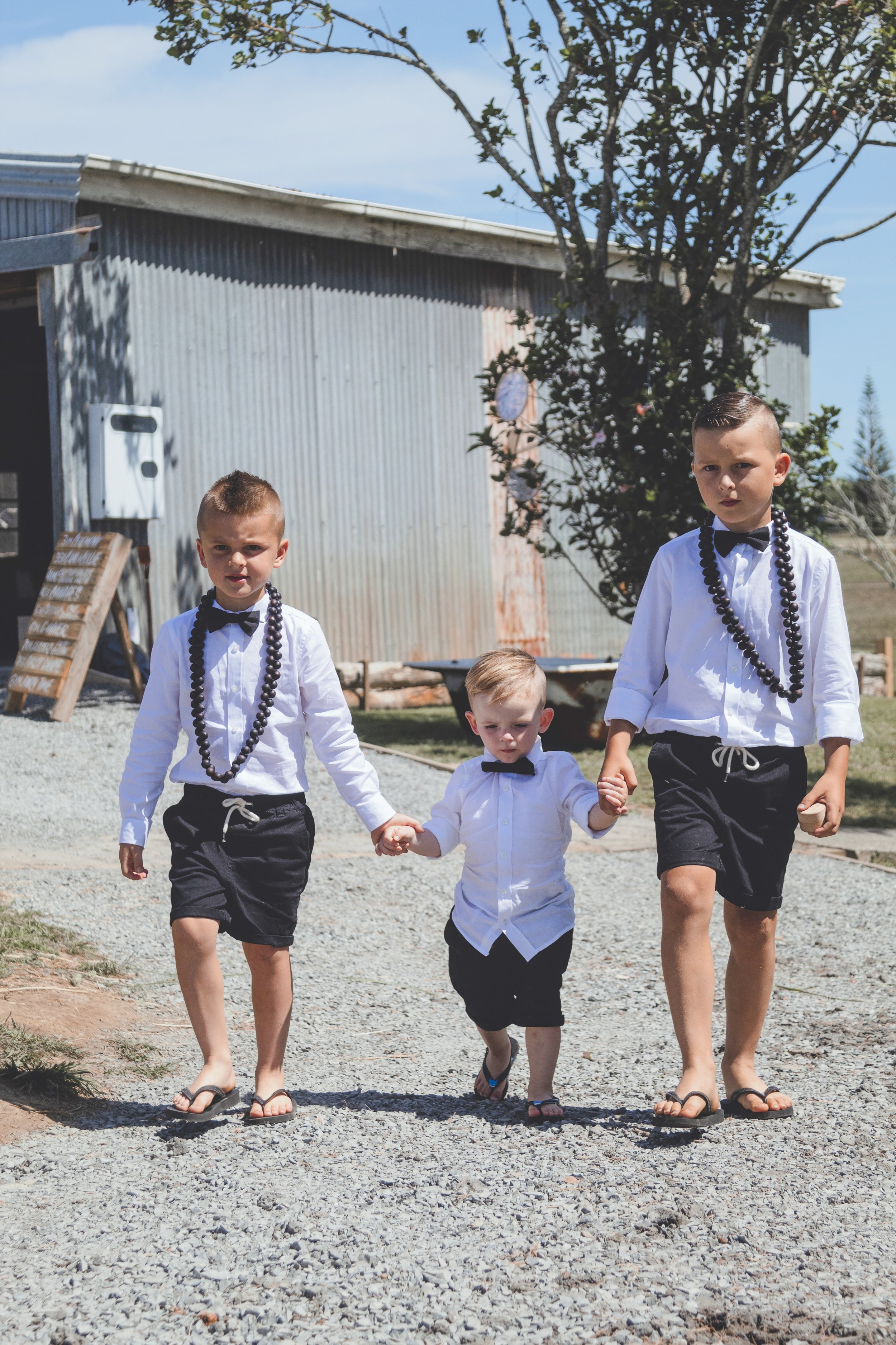 Three boys in white shirts and black shorts holding hands, walking on a gravel path outdoors, dressed in formal attire with beads and bow ties, under a clear sky with a metal building and trees in the background.