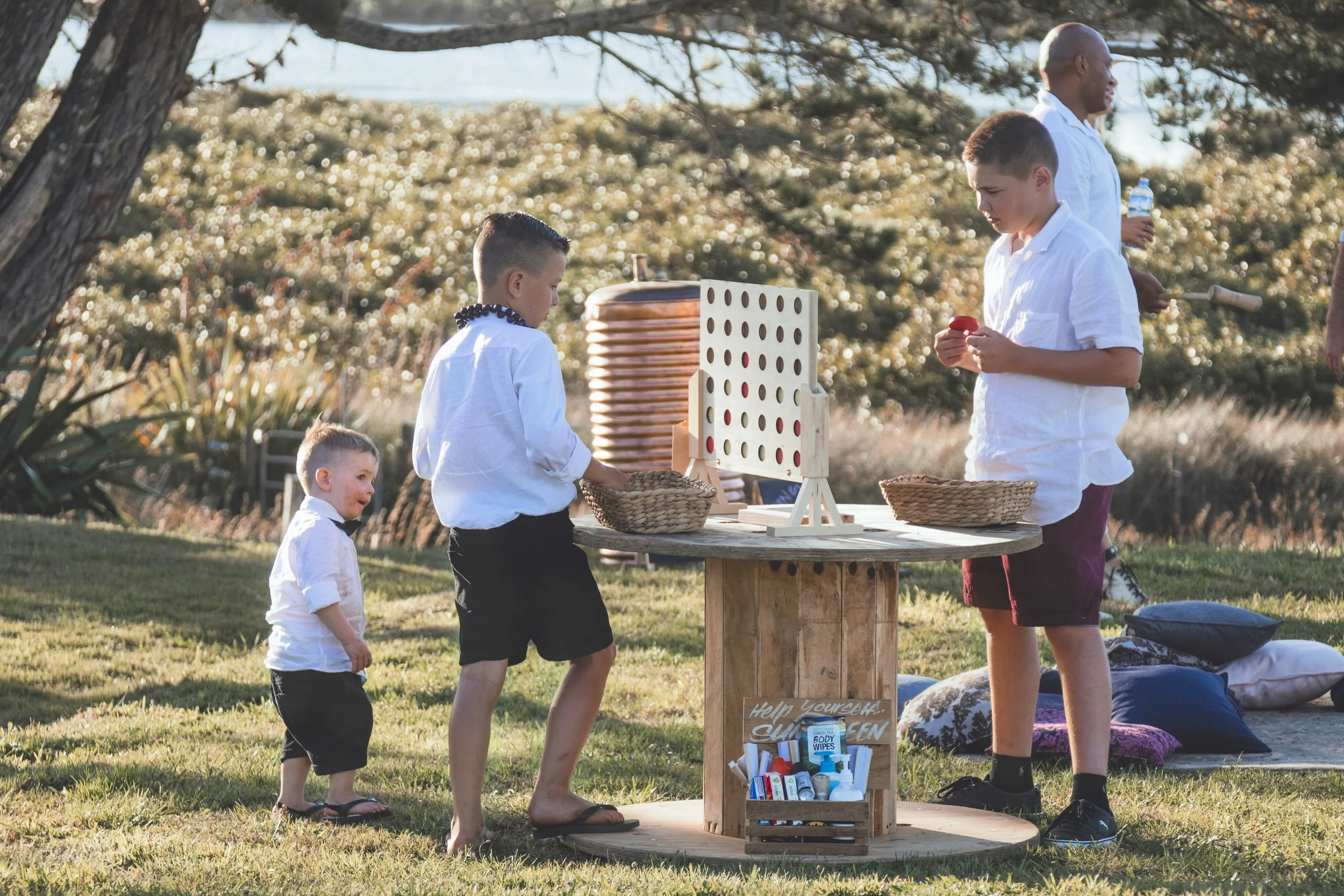 A group of children and an adult playing Connect Four outdoors near a tree and a body of water, with sunlight shining through the branches.