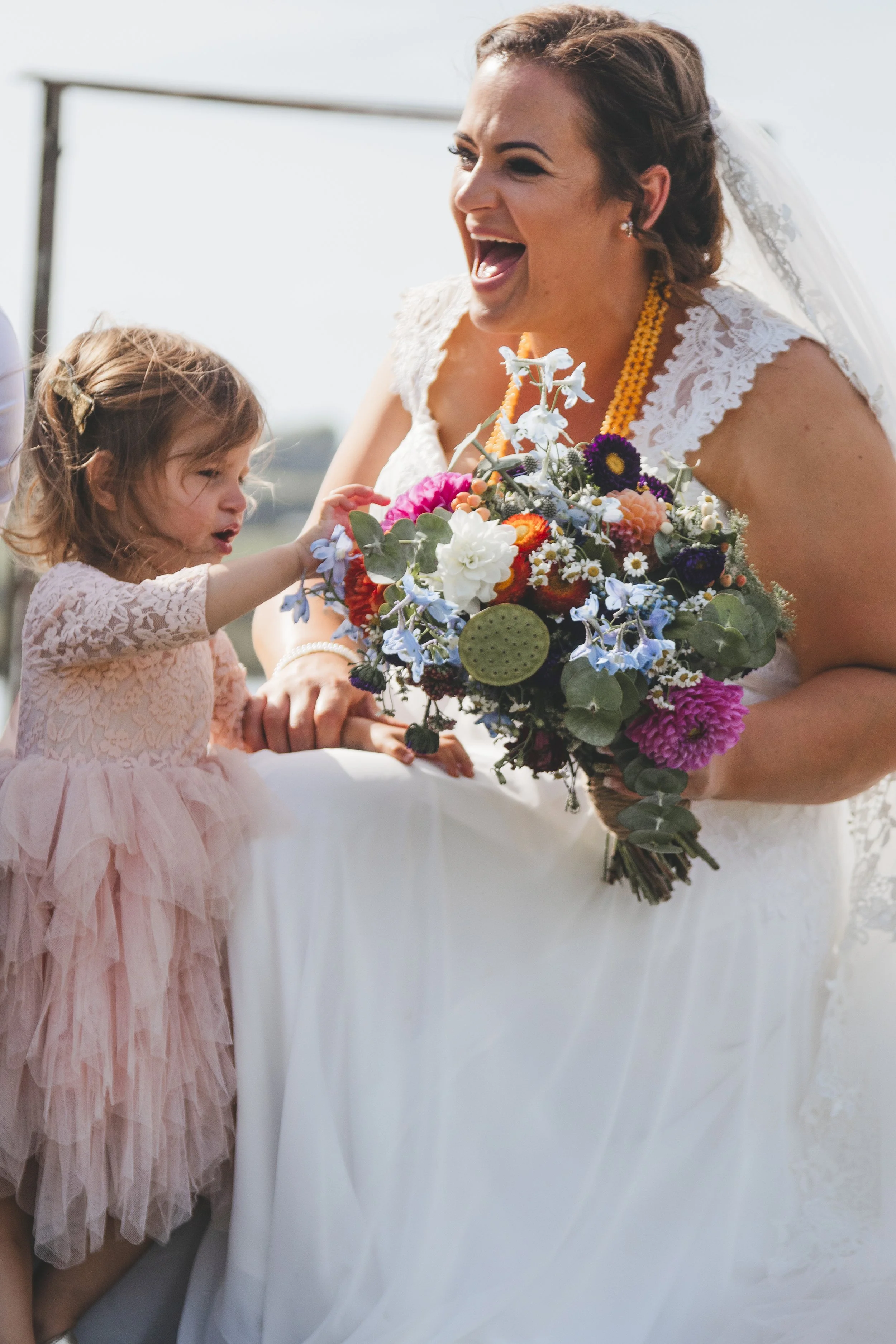 A woman smiling and holding a bouquet of colorful flowers while wearing a wedding dress and veil, with a young girl in a pink dress touching the bouquet.
