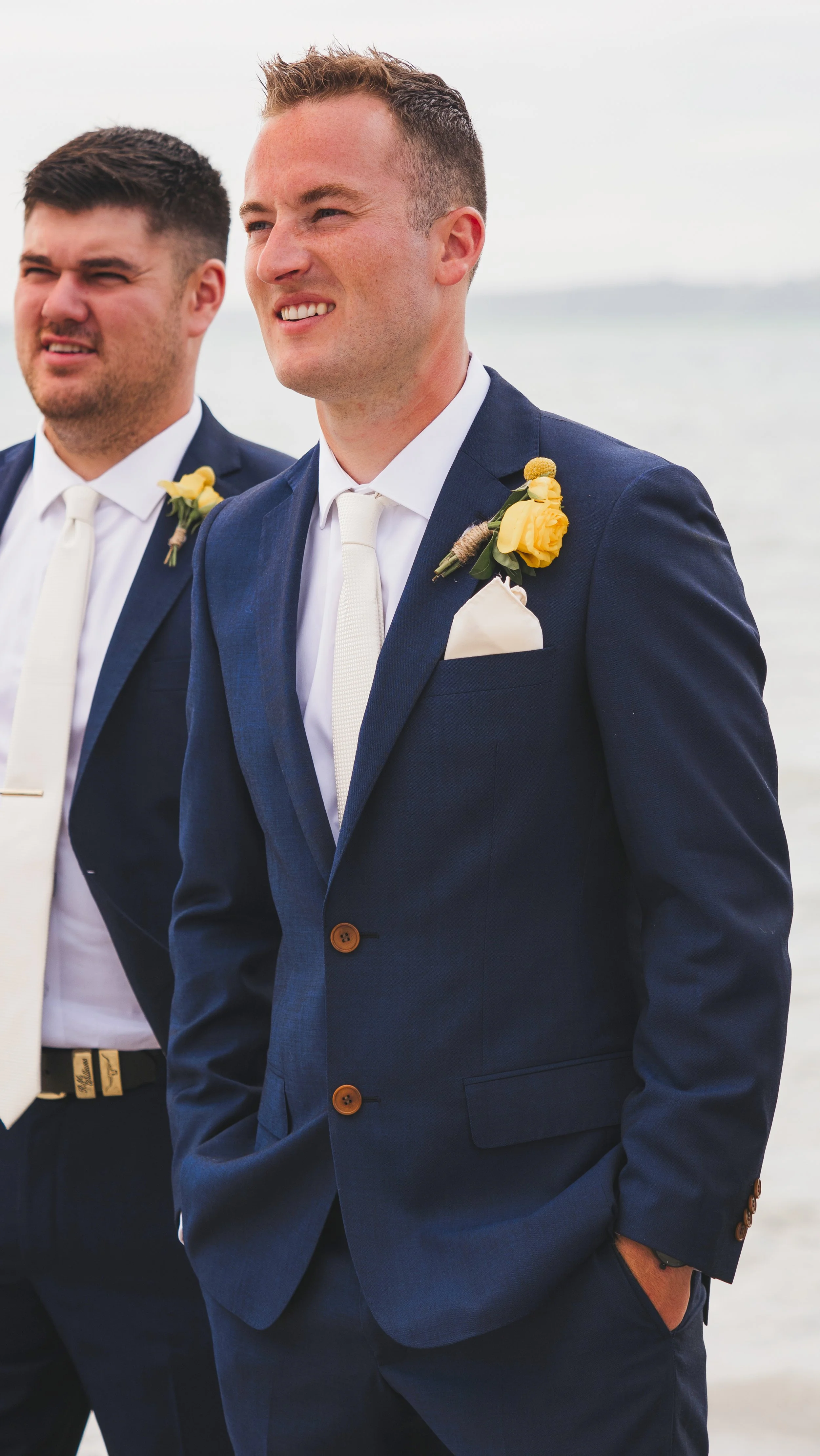 Two men in navy suits with yellow boutonnieres, white shirts, and ties, standing outdoors near water with a blurred shoreline in the background.