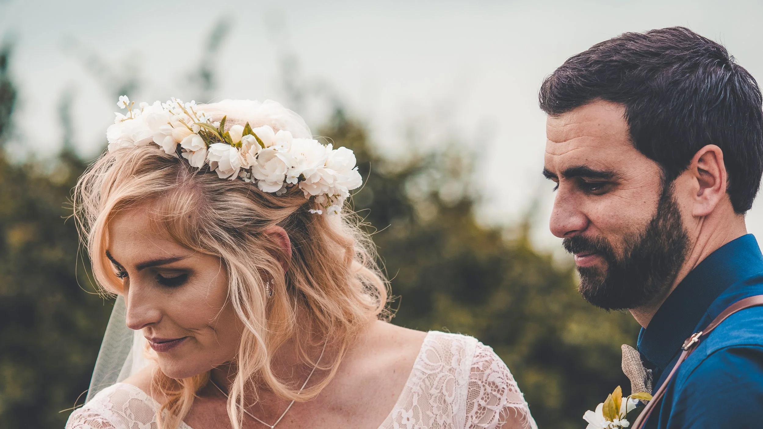 A bride with blonde hair and a floral crown and a groom with dark hair and a beard standing close together outdoors.