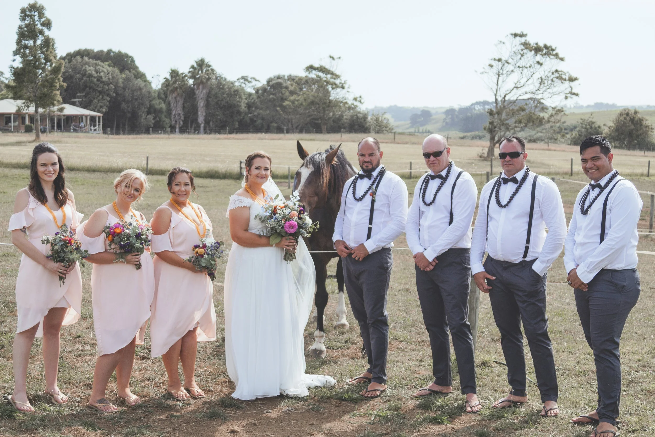 A wedding party standing outdoors on a grassy field, with women in light pink dresses holding bouquets, and men in white shirts, dark trousers, suspenders, bow ties, and sunglasses, with a bride in a white dress holding a bouquet, and a groom with a 